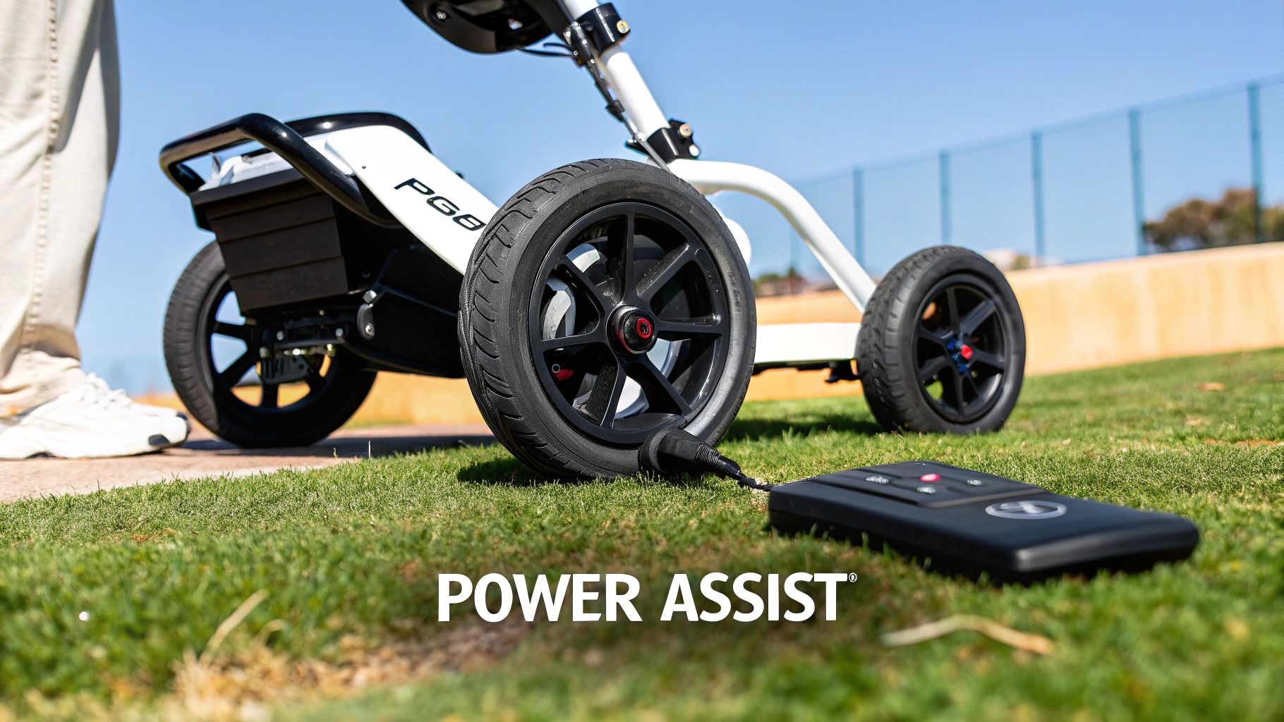 A white electric golf push cart with black wheels and a remote control on green grass, emphasizing 'POWER ASSIST'.