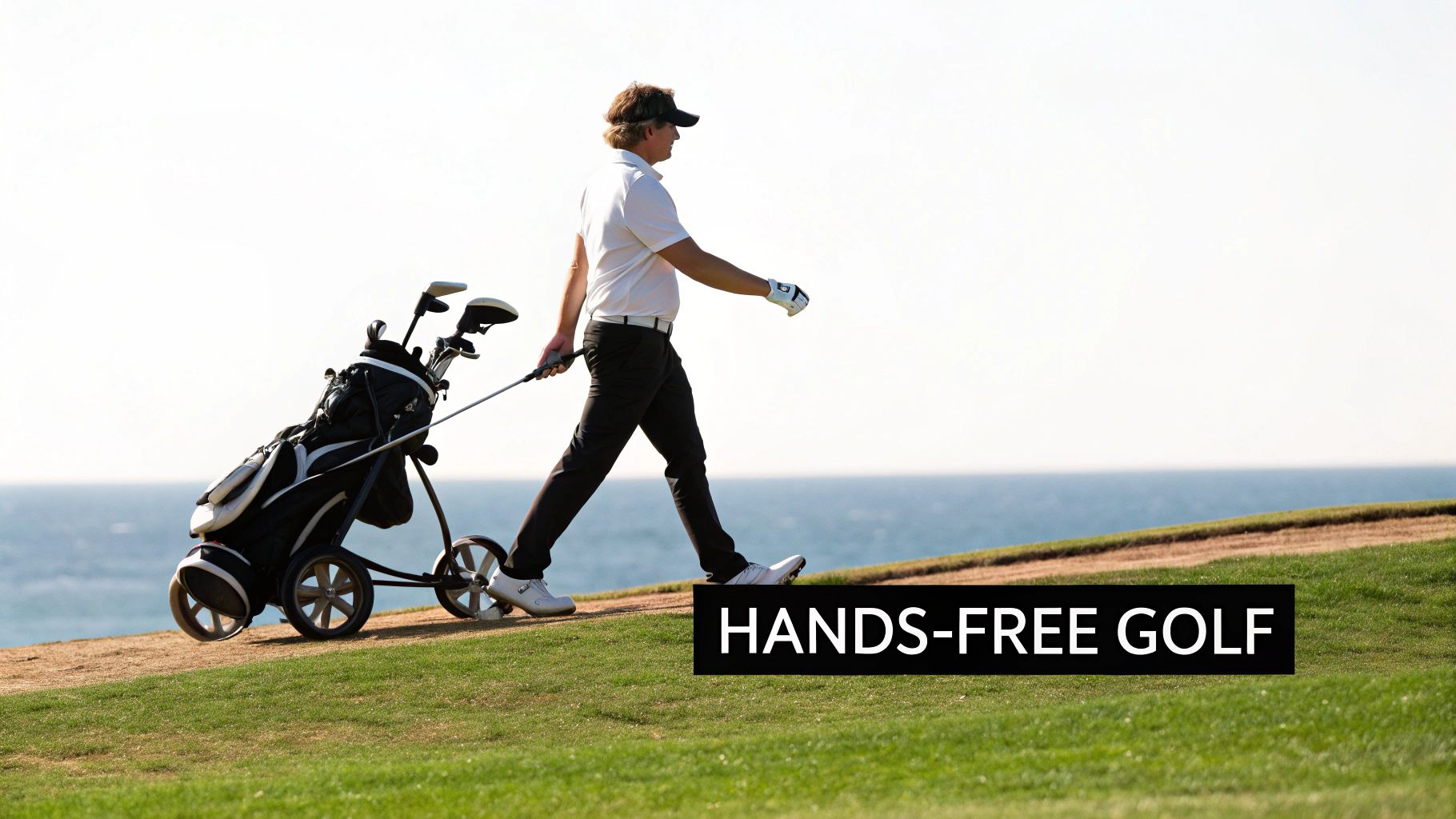 A male golfer walks on a green course, pulling a golf bag trolley with the ocean beyond.