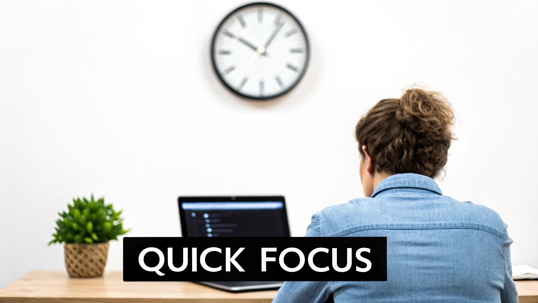 Person from behind works on a laptop at a desk with a plant and clock on a white wall.