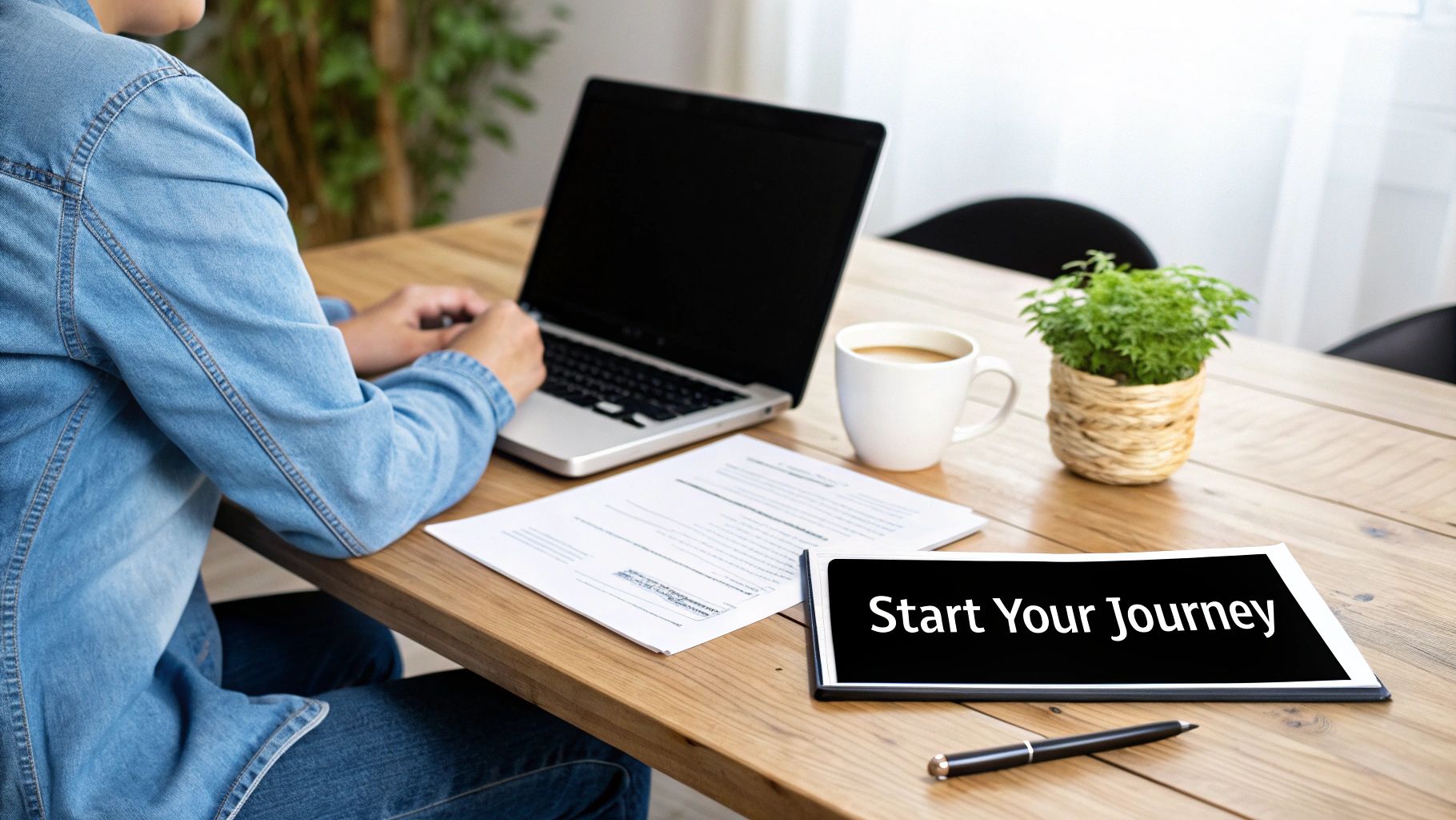 A person in a denim shirt typing on a laptop with papers, coffee, and a 'Start Your Journey' sign.