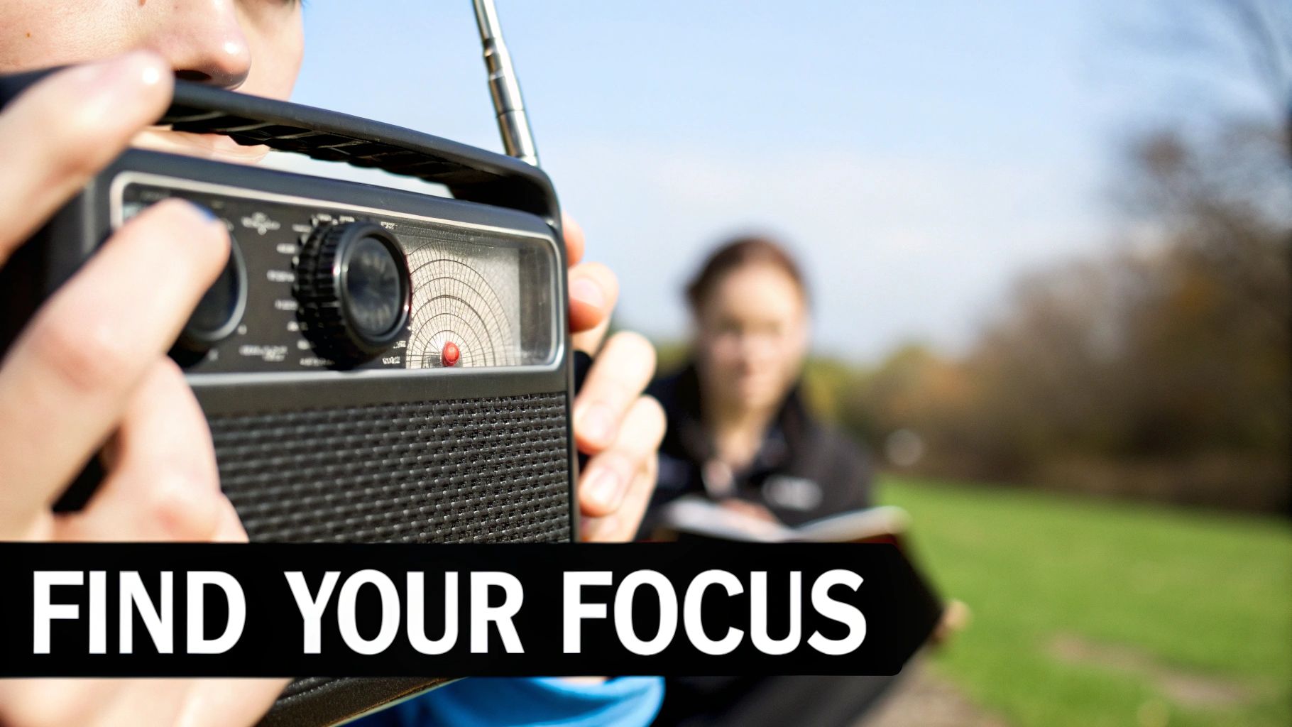 Hands turning a knob on a vintage portable radio outdoors, with a person reading in the background.
