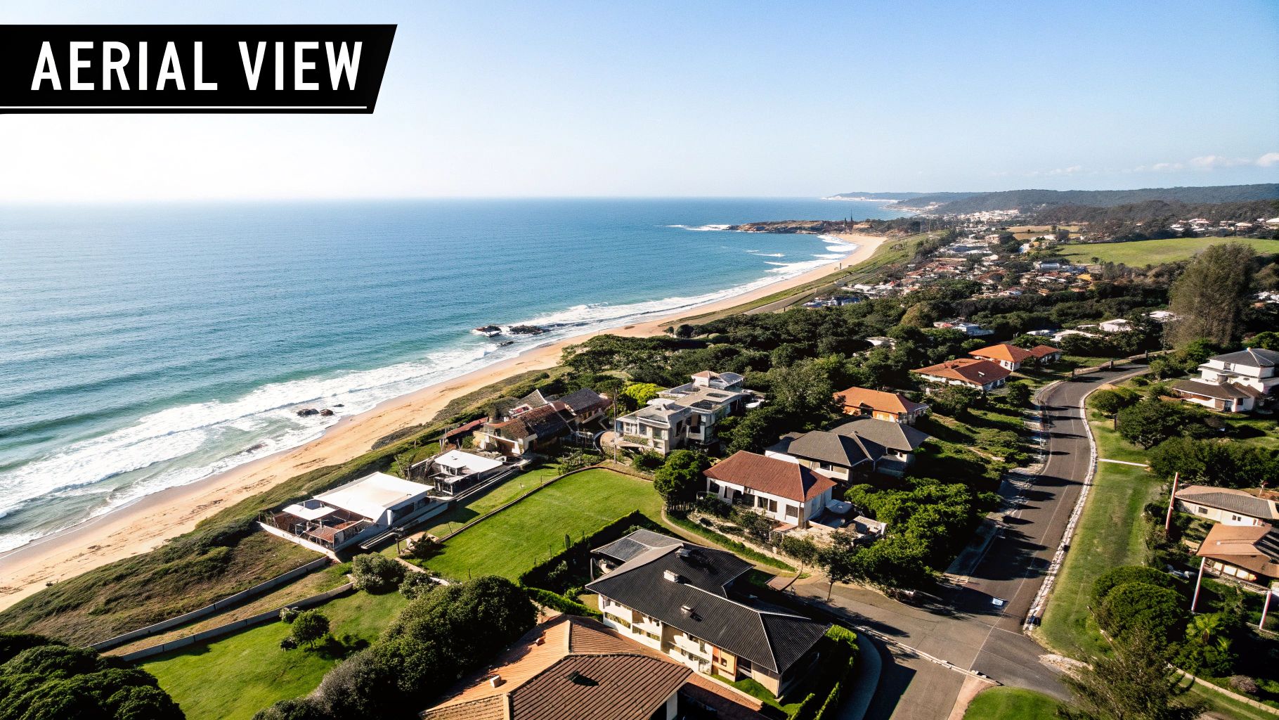 An aerial view of a scenic coastline featuring a sandy beach, blue ocean, and residential homes.