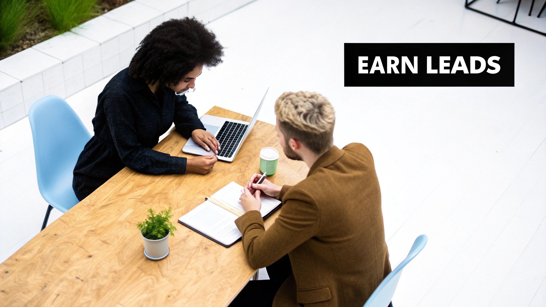 Two colleagues collaborate at a desk, one uses a laptop while the other writes, promoting lead generation.