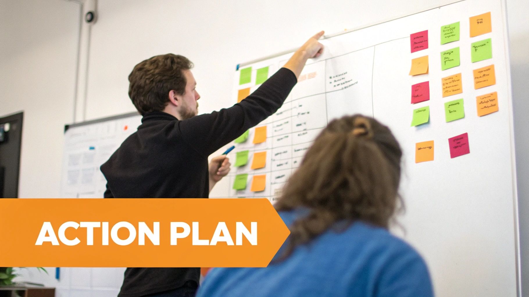 A man points at a whiteboard with colorful sticky notes, explaining an action plan to a colleague.