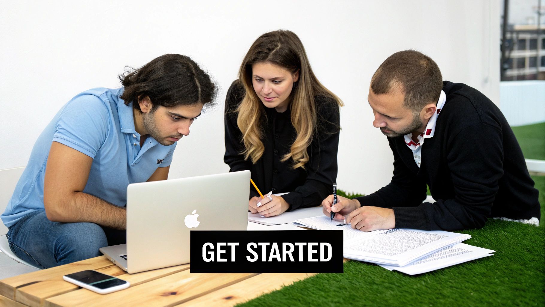 Three young professionals collaborate around a laptop and notebooks on a modern workspace setup.