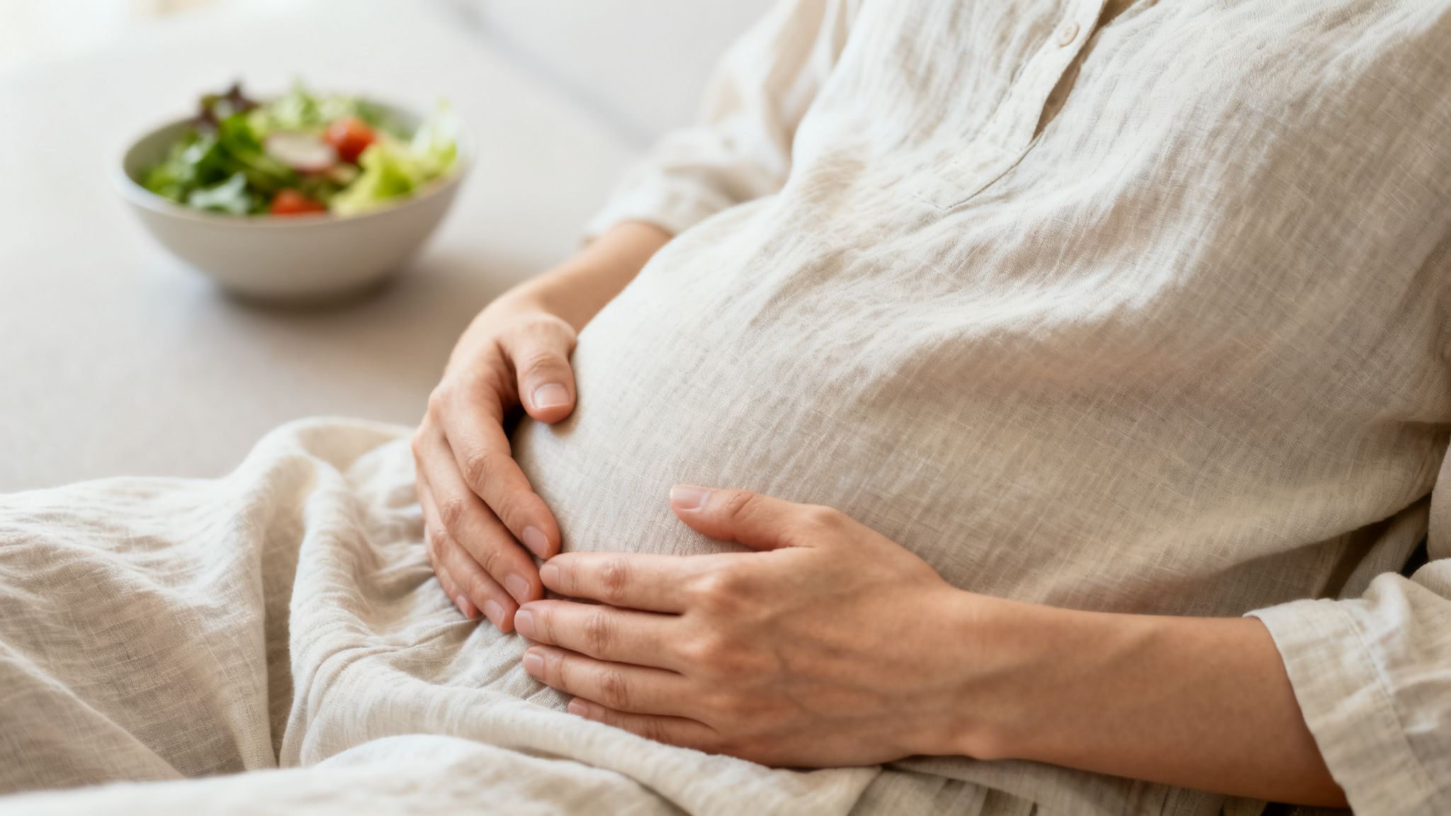 A woman's hands gently rest on her stomach, with a healthy salad on the table in front of her.