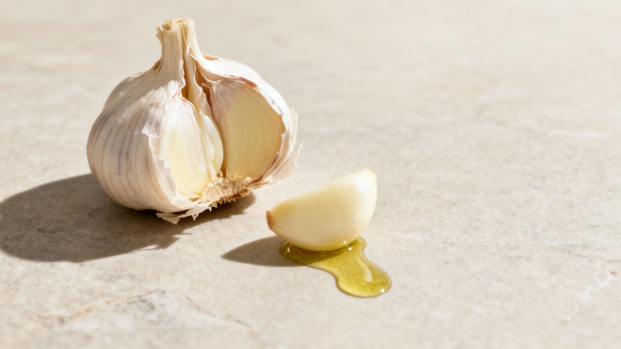 A partially opened garlic bulb and a single peeled clove resting in a small pool of oil on a light counter.