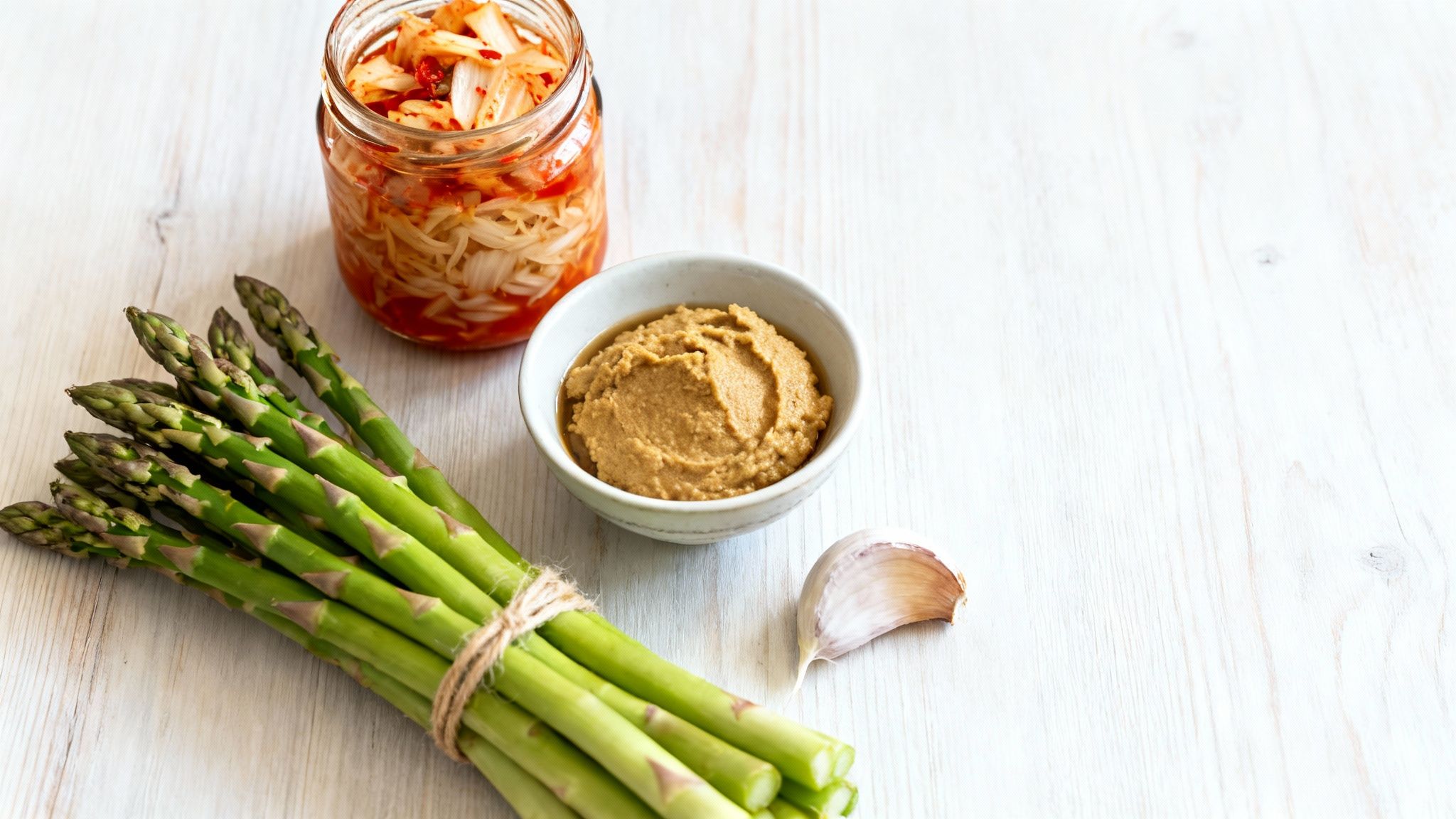 A jar of kimchi, a bowl of miso paste, fresh asparagus, and garlic on a light wood table.