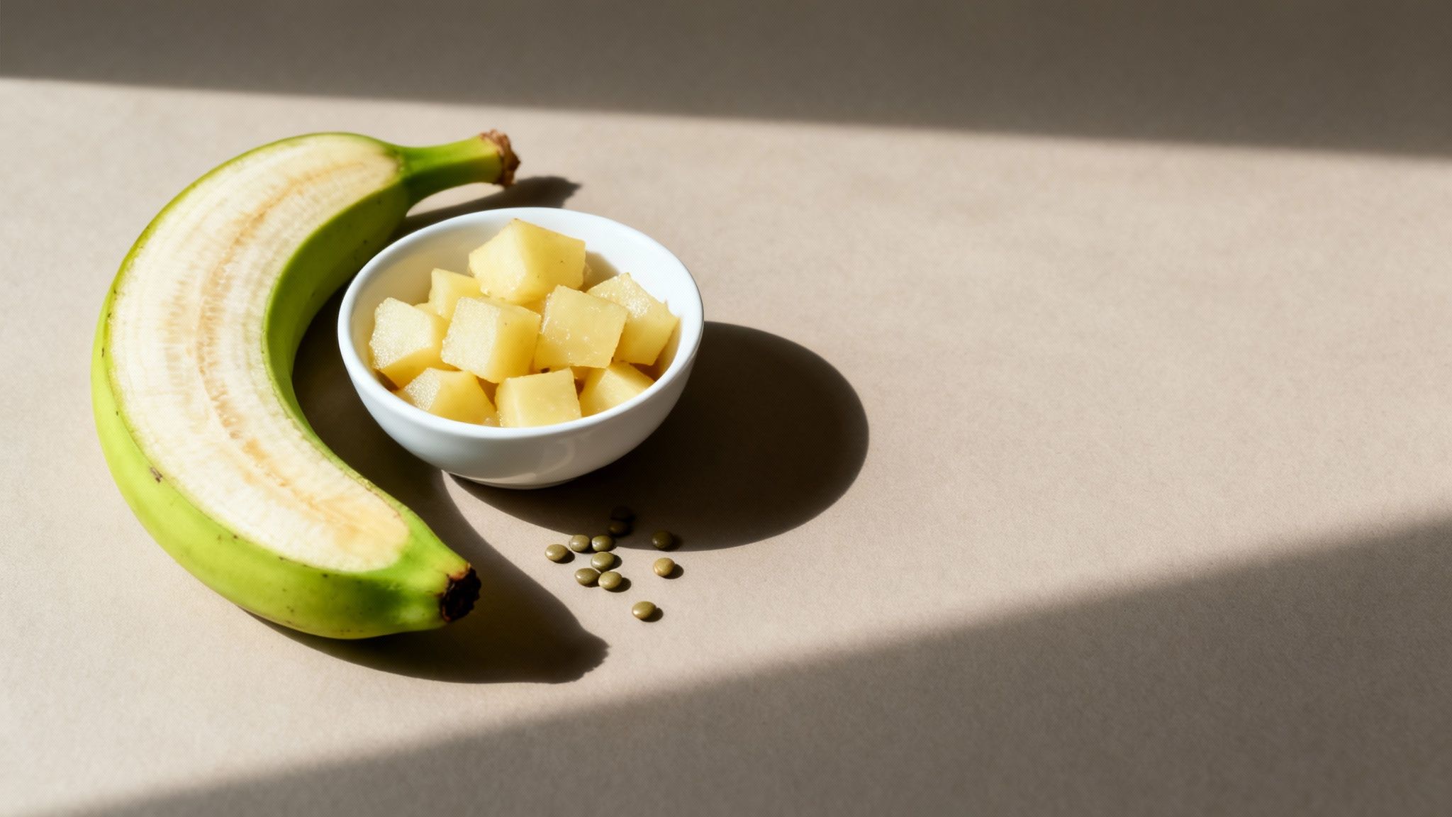 A sliced green banana, a bowl of diced tropical fruit, and lentils on a brown surface with shadows.