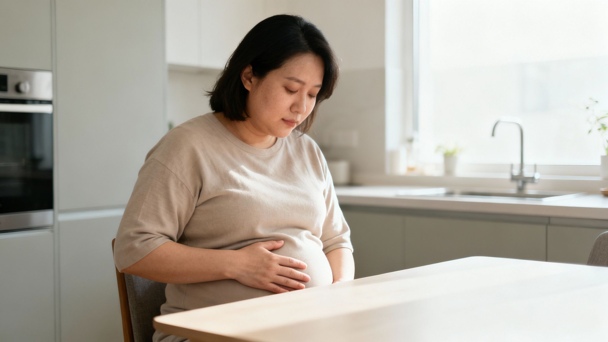 Middle-aged Asian woman experiencing digestive discomfort while sitting at kitchen table