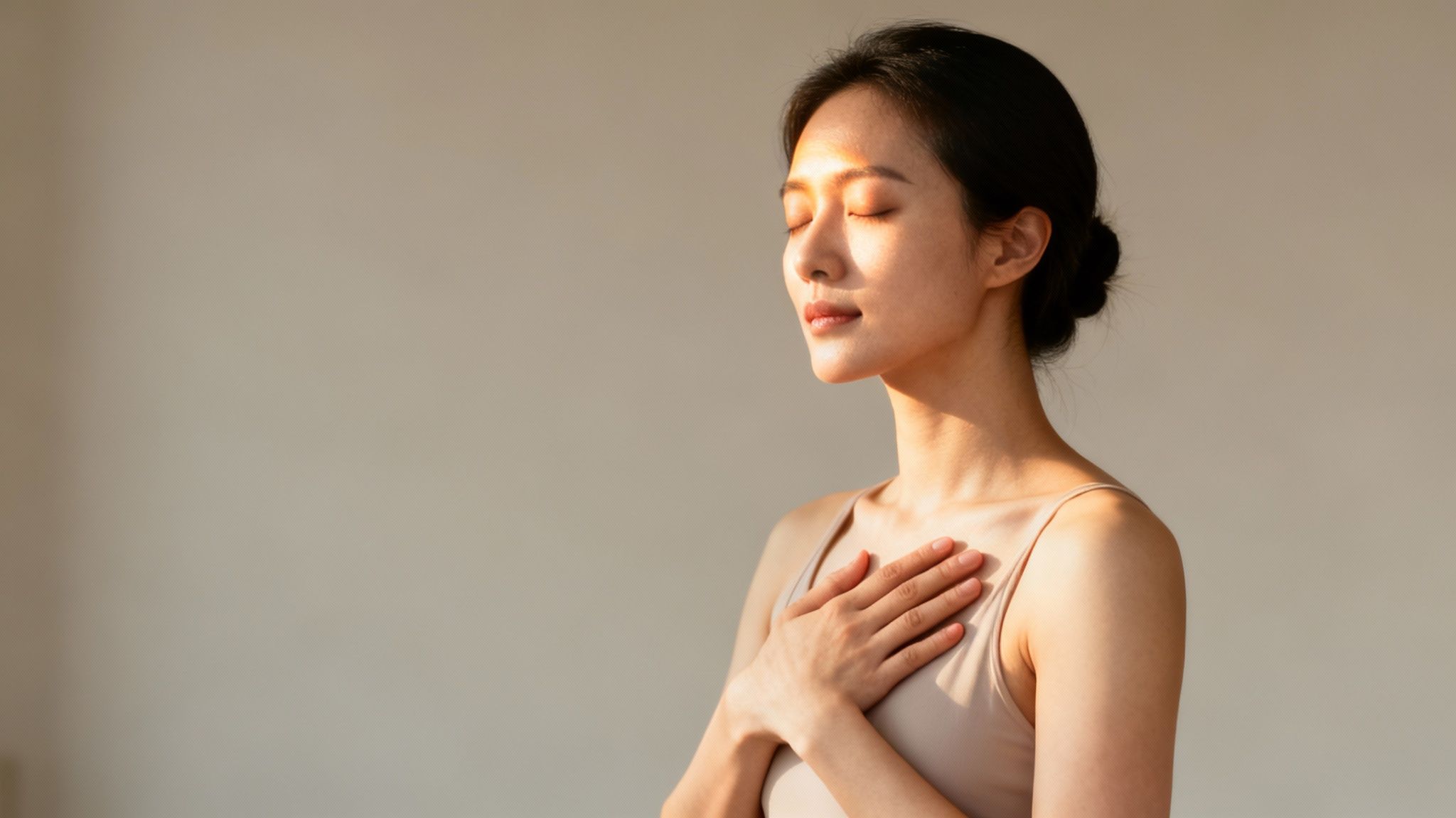 Serene Asian woman meditating with closed eyes and hand on chest, bathed in warm sunlight.