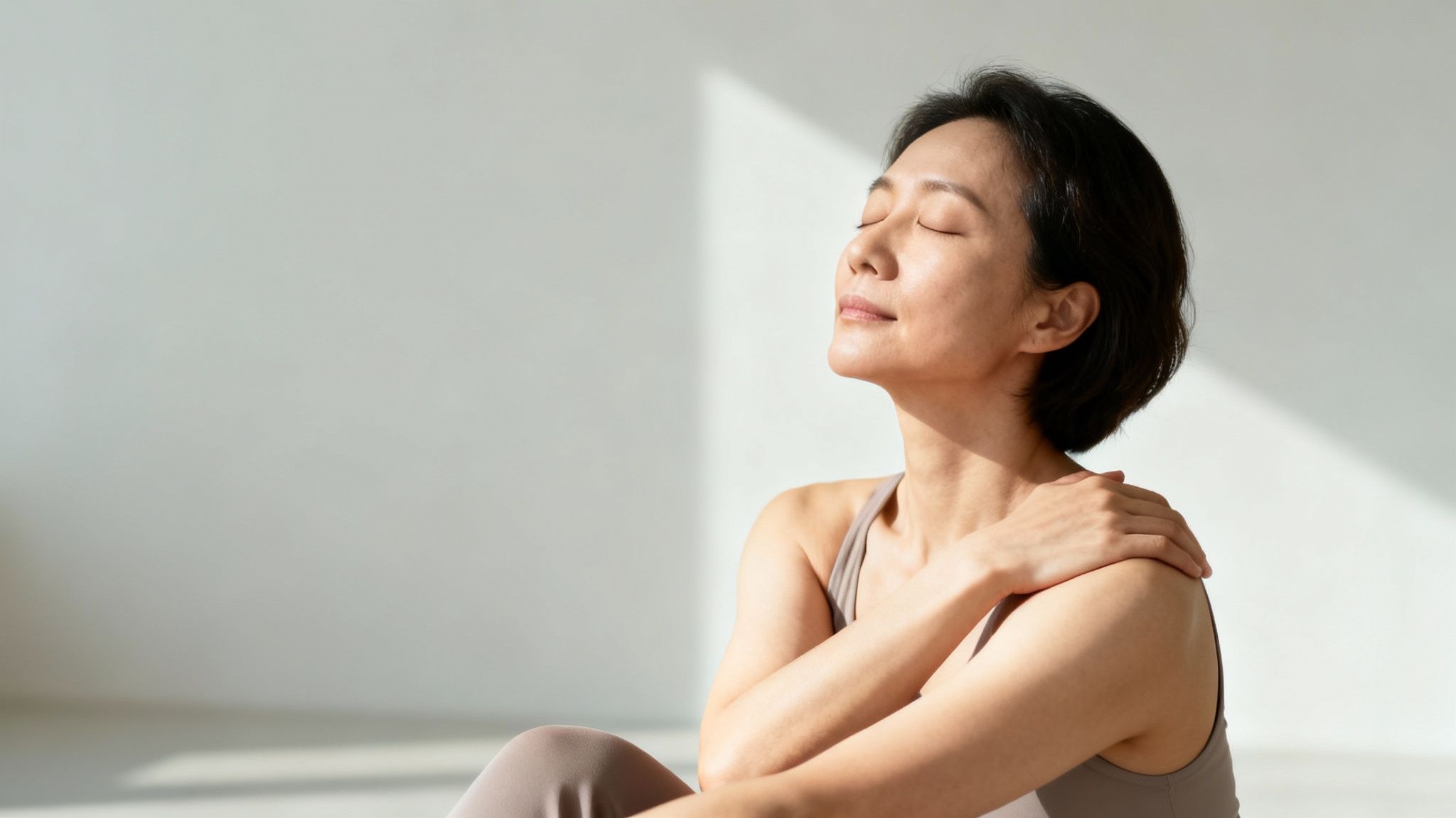 A serene woman with closed eyes enjoys sunlight, her hand resting on her shoulder.