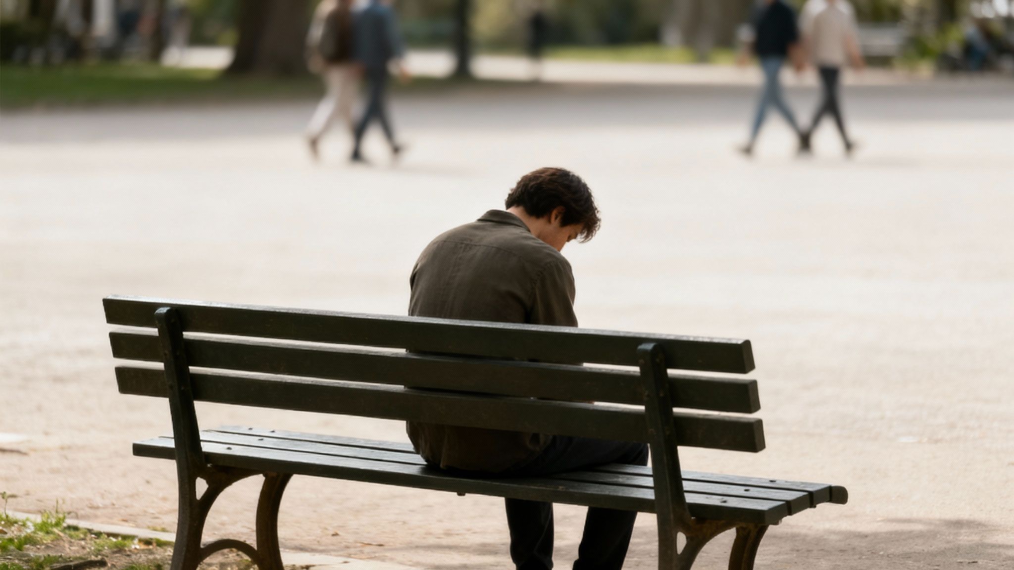 A person sits alone on a dark green park bench, looking down, with blurred people walking in the background.