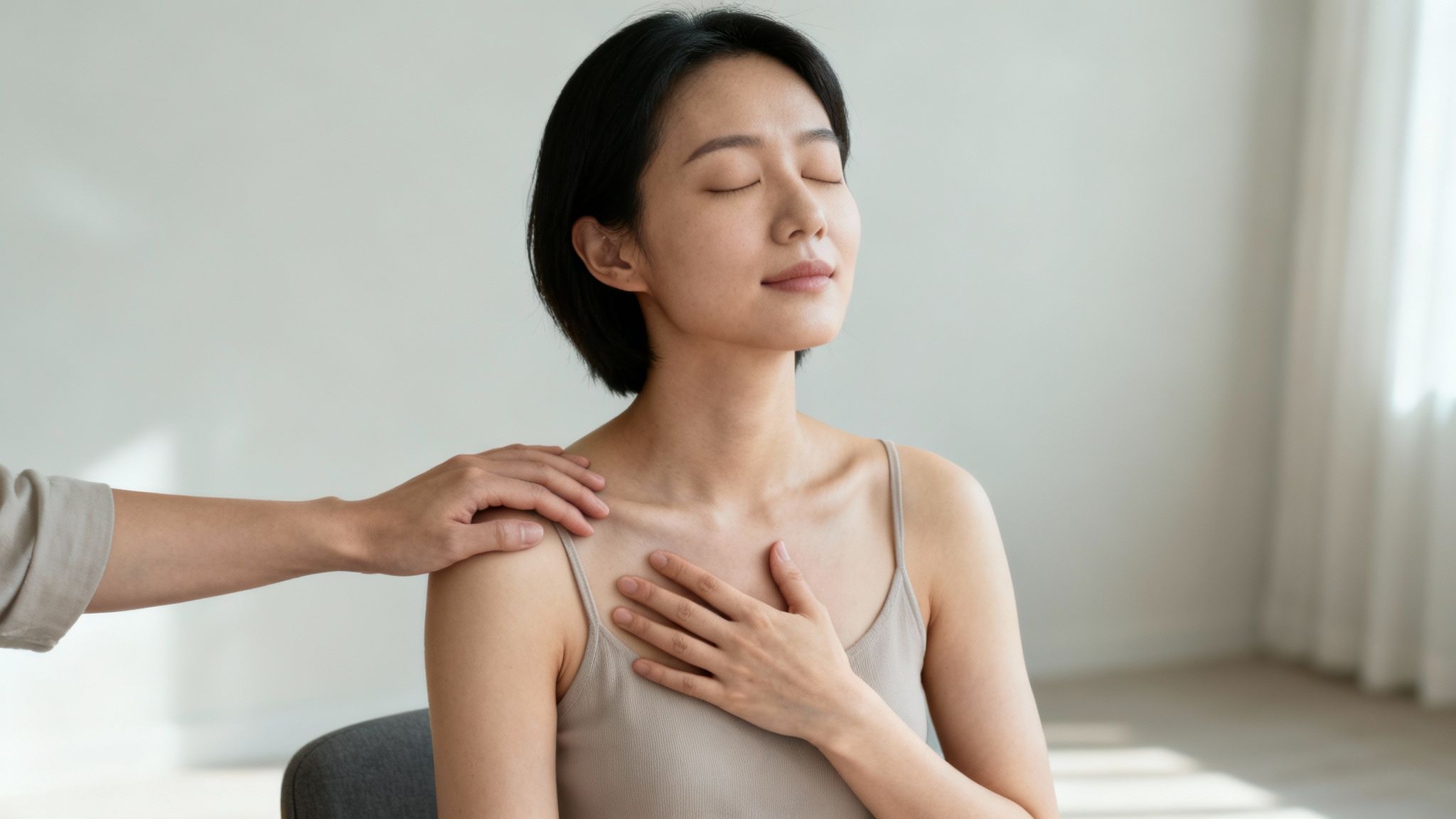 A serene woman with closed eyes is gently supported by a hand on her shoulder and her own hand on her chest.