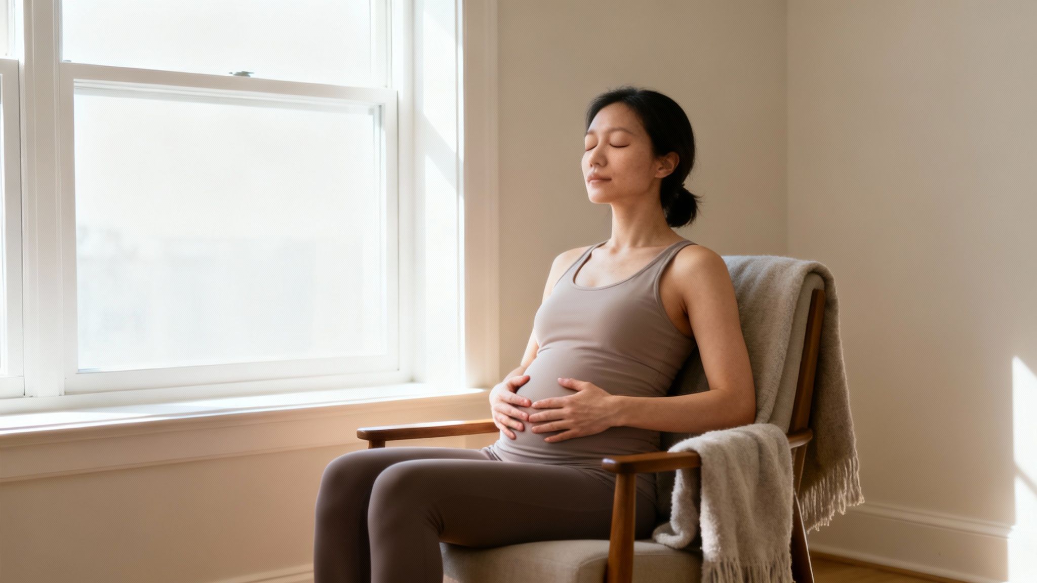 Pregnant woman peacefully meditating while sitting in chair by sunny window at home