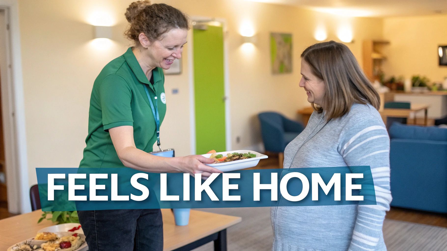 A friendly caregiver in a green uniform serving a healthy meal to a smiling woman in a home-like setting.