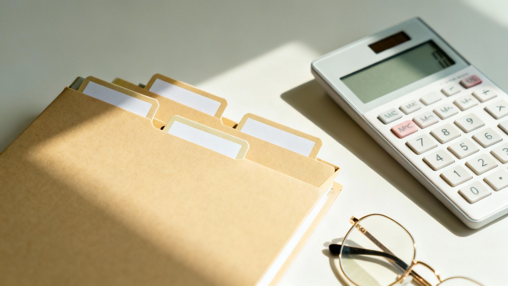 밝은 햇살 아래 서류 파일, 계산기, 안경이 놓인 사무실 책상 풍경.