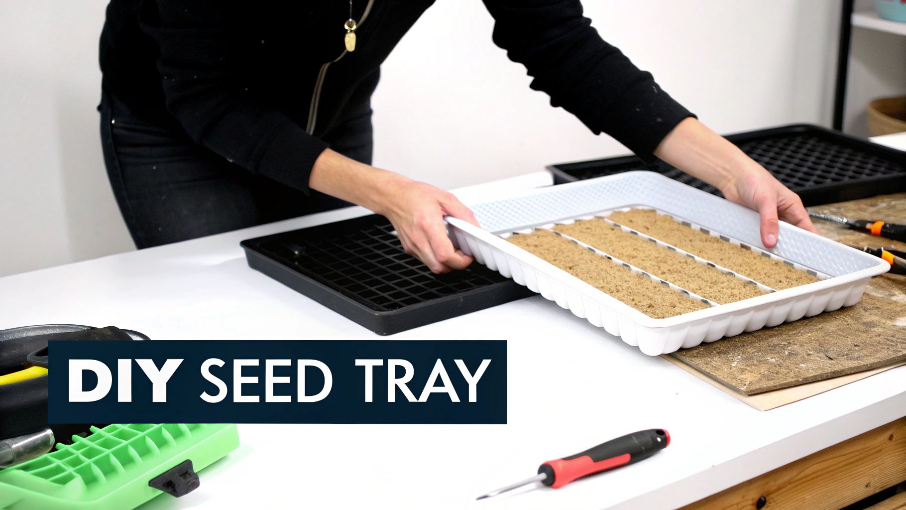 A person's hands are carefully placing a white DIY seed tray filled with soil into a black grid tray on a white table.