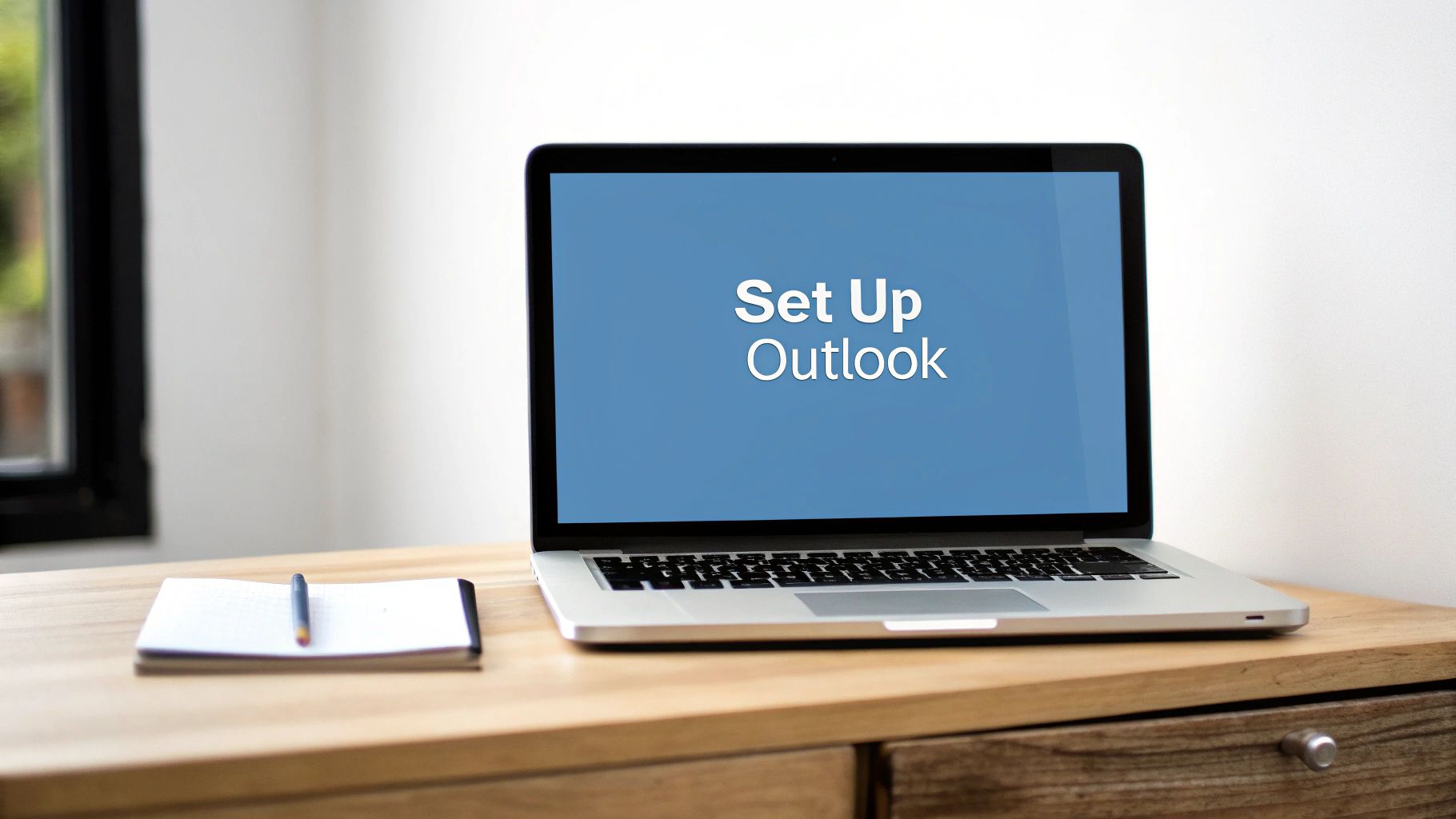 A silver laptop displaying "Set Up Outlook" on its blue screen, placed on a wooden desk with a notebook and pen.
