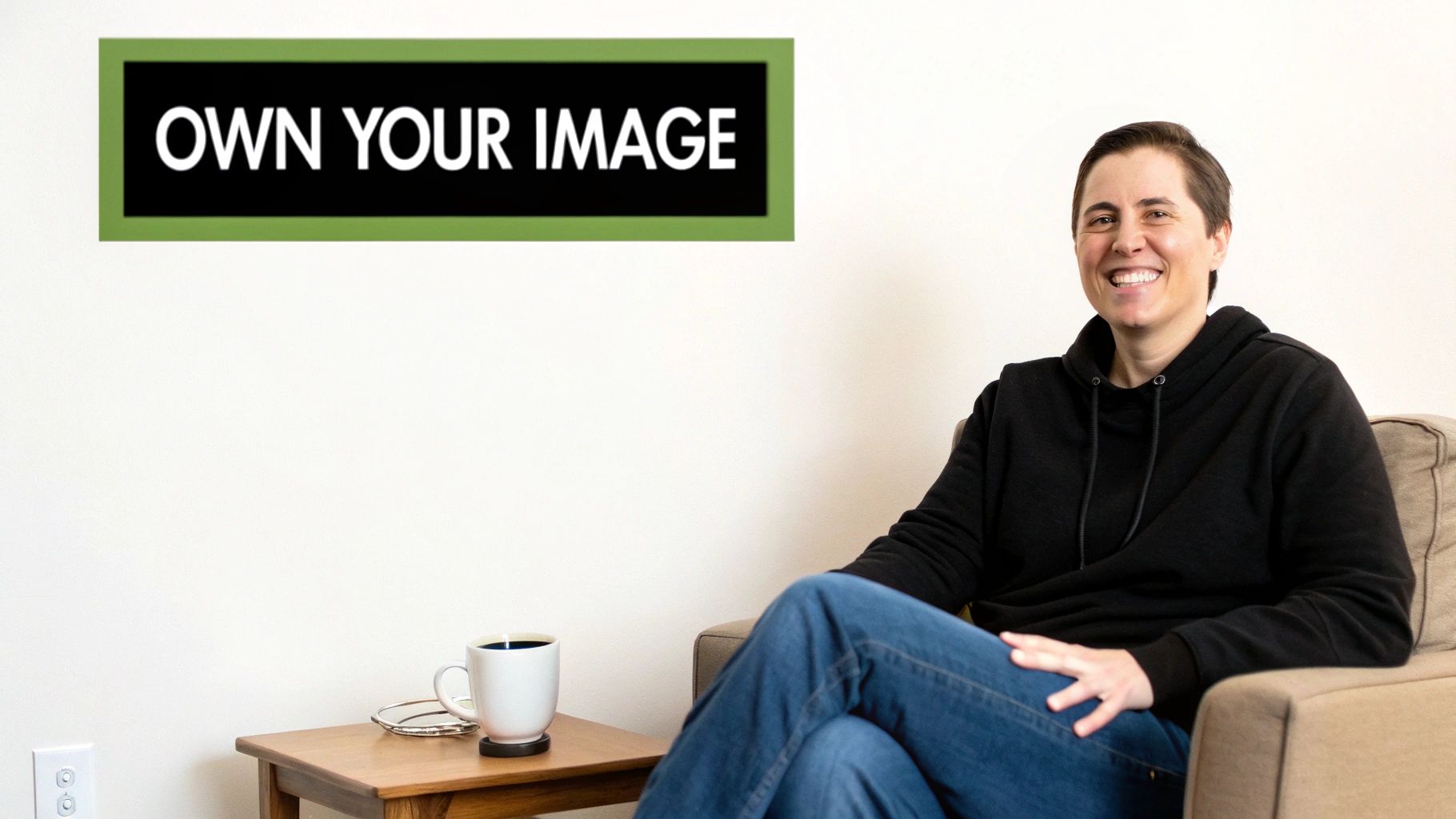 A smiling person in a black hoodie sits on a couch, next to a sign that says 'OWN YOUR IMAGE'.