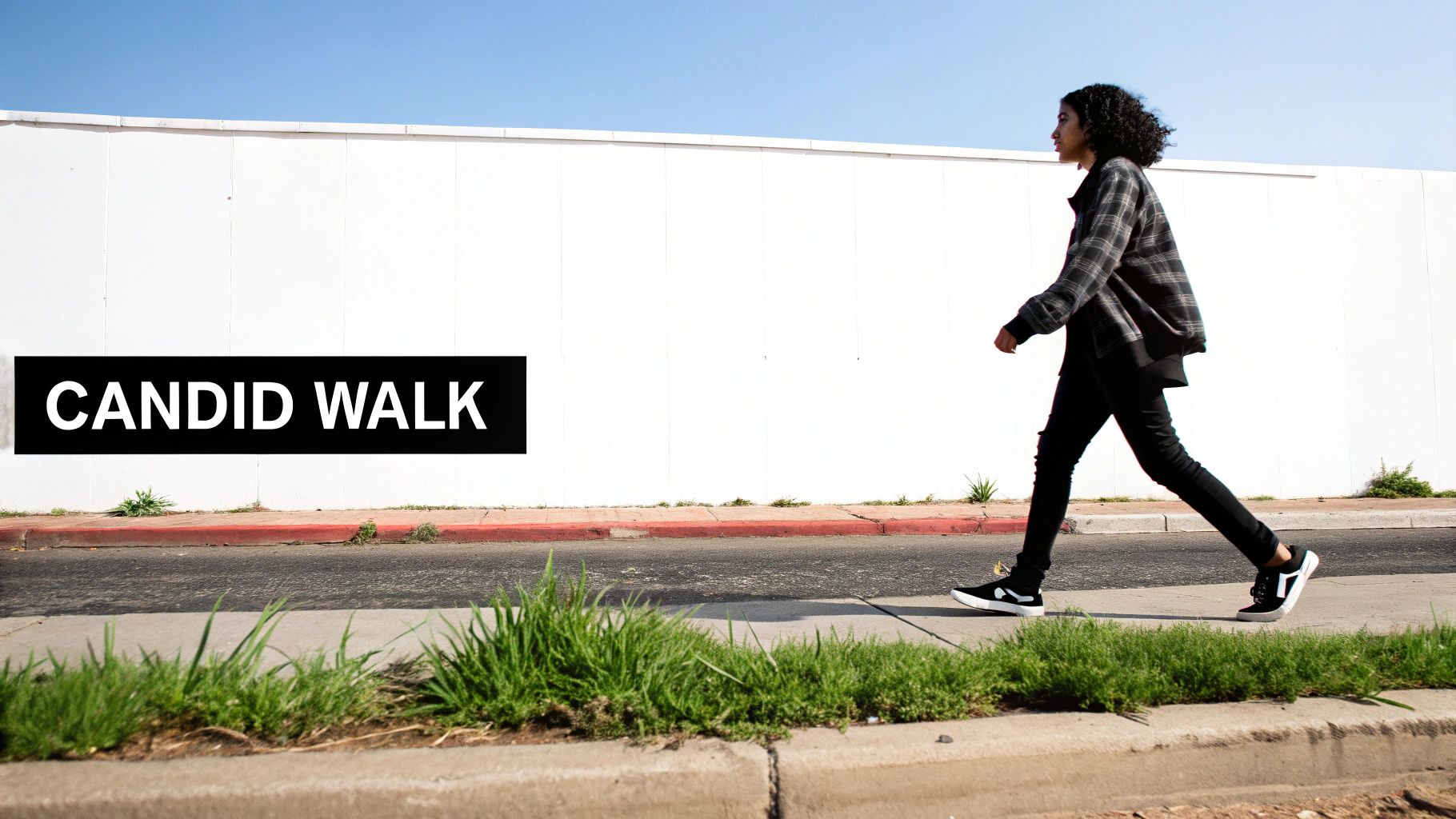 A person with curly hair walks casually on a sidewalk next to a white wall with 'CANDID WALK' text.