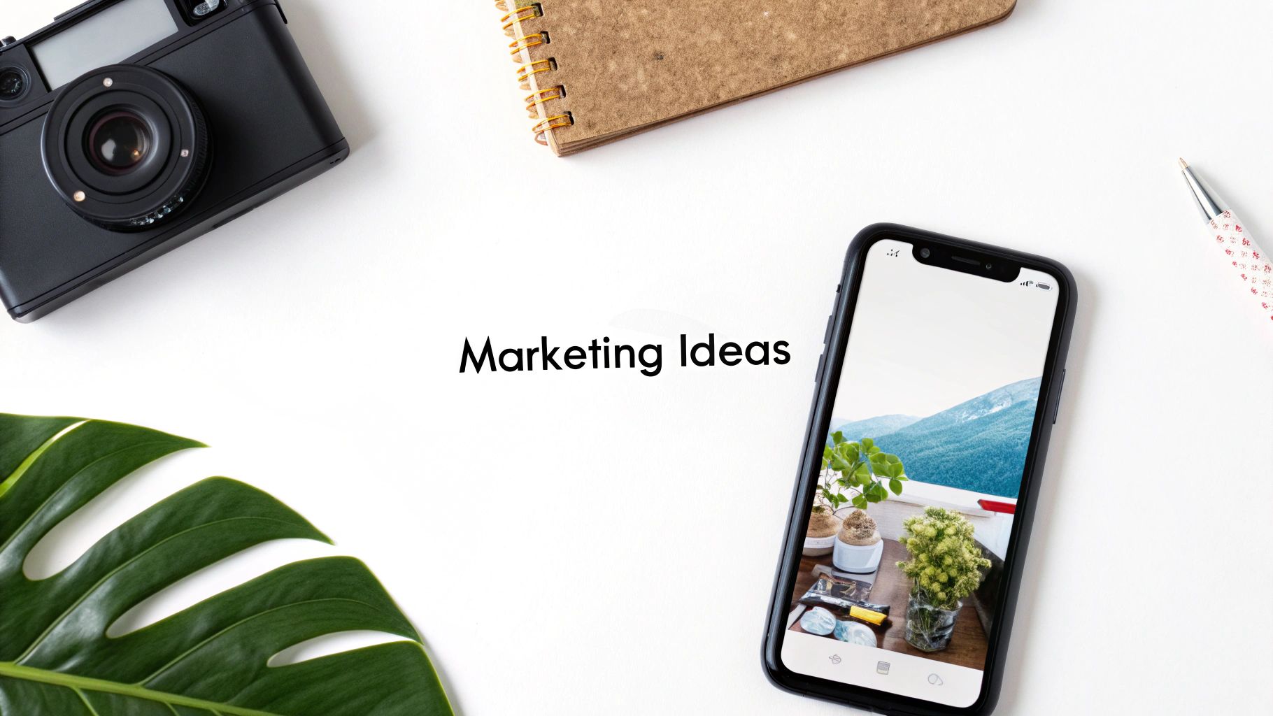 Flat lay of a camera, notebook, smartphone showing a scenic view, pen, and Monstera leaf, with 'Marketing Ideas' text.