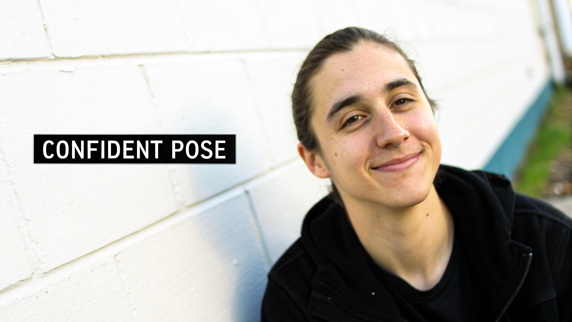 Eine junge Person mit zurückgebundenem dunklem Haar, die selbstbewusst lächelt und ein schwarzes Oberteil vor einer weißen Backsteinwand trägt.