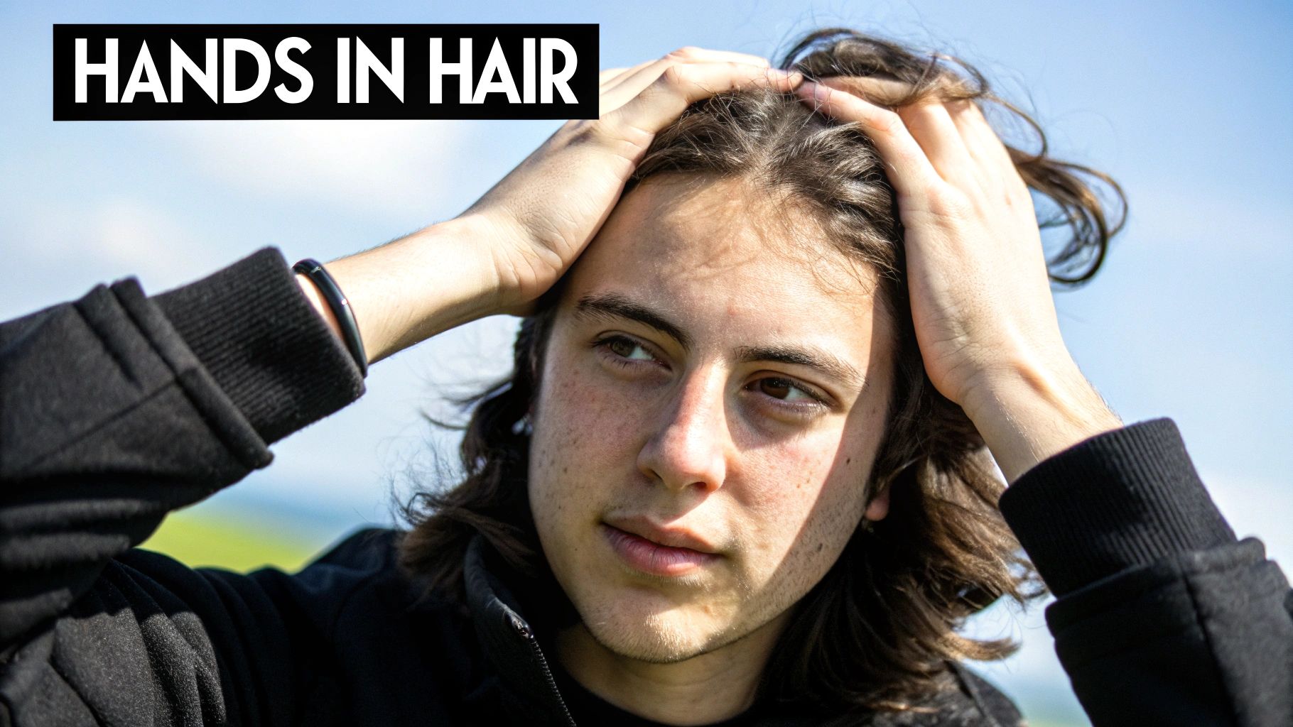 A young person with dark, wavy hair holds their hands on their head, looking to the side under a blue sky.