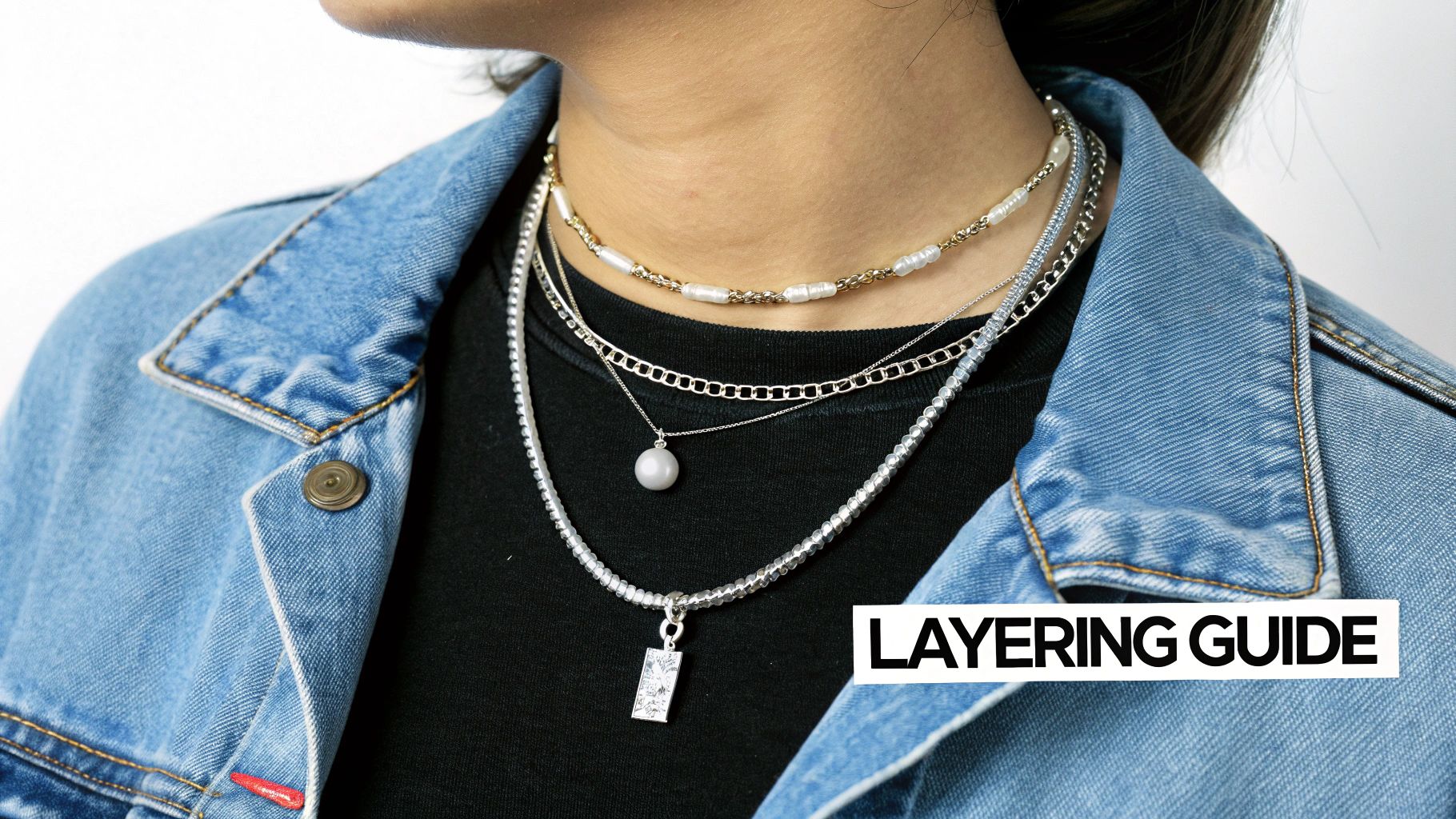 A close-up of a person wearing a denim jacket and multiple layered necklaces, featuring pearls and silver chains.