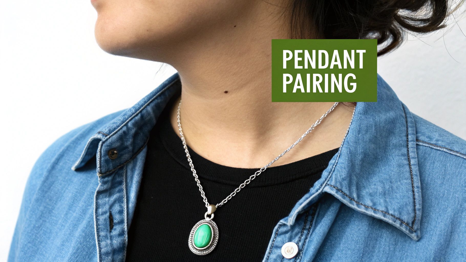 Close-up of a person wearing a silver chain with a green oval pendant over a black t-shirt and denim shirt.