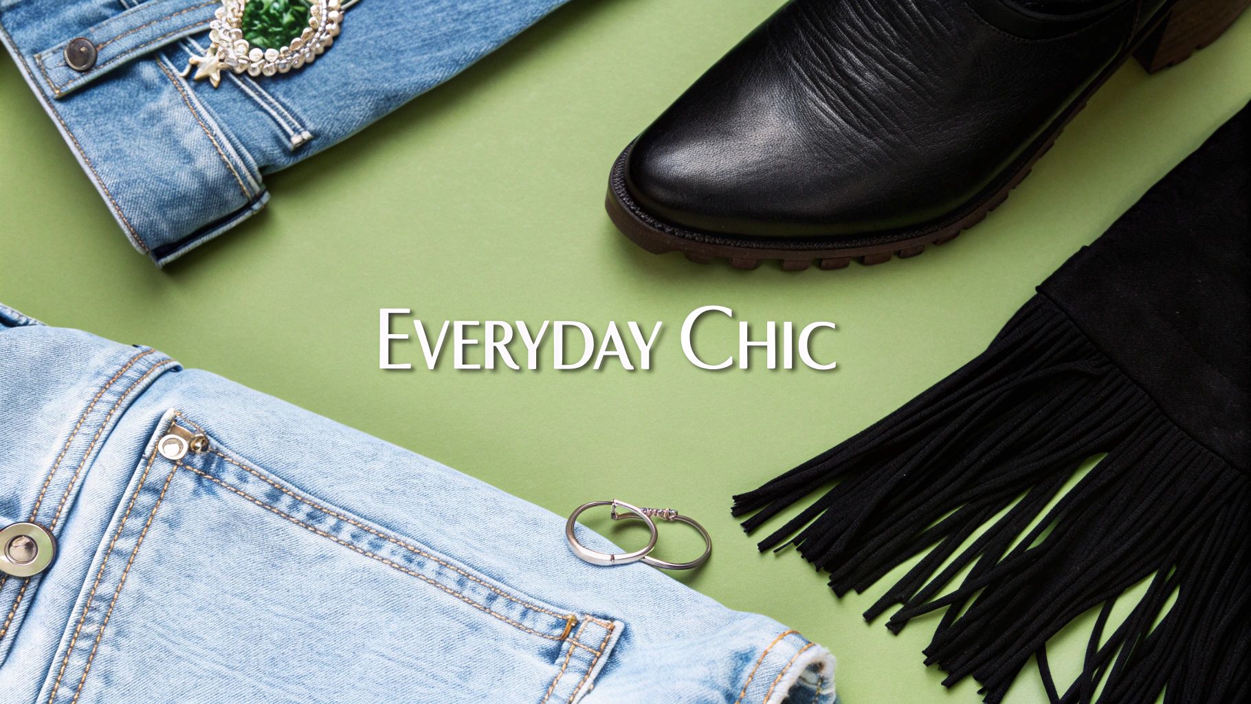 A flat lay of everyday chic fashion items including a denim jacket, black boot, fringed fabric, and silver rings on a green background.