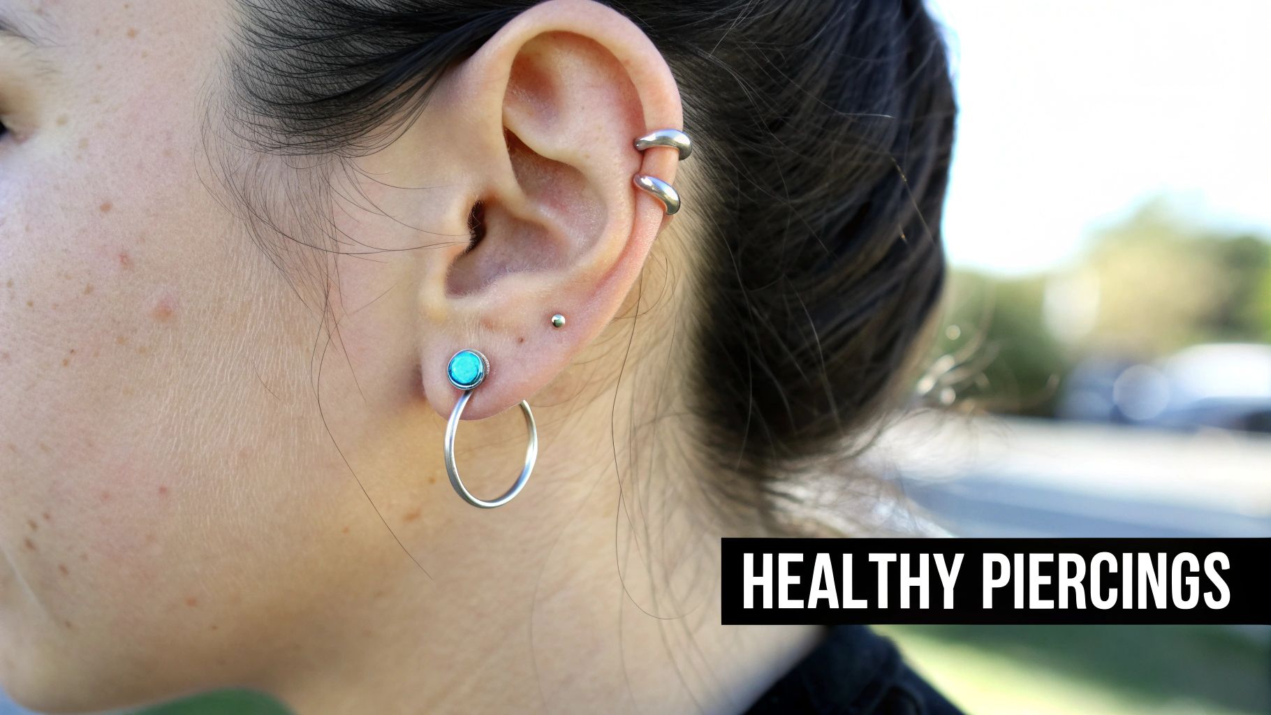 Close-up of a person's ear with multiple healthy piercings, including a teal stud, silver hoop, and helix cuffs.