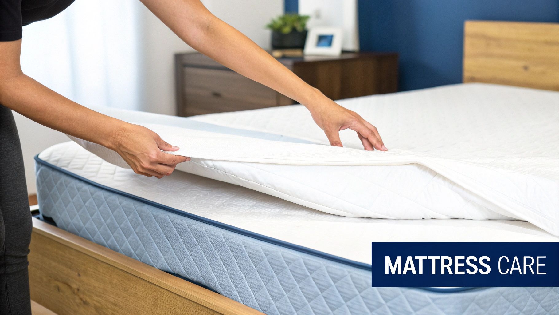 A person's hands adjust a white mattress protector over a blue quilted mattress in a bedroom.