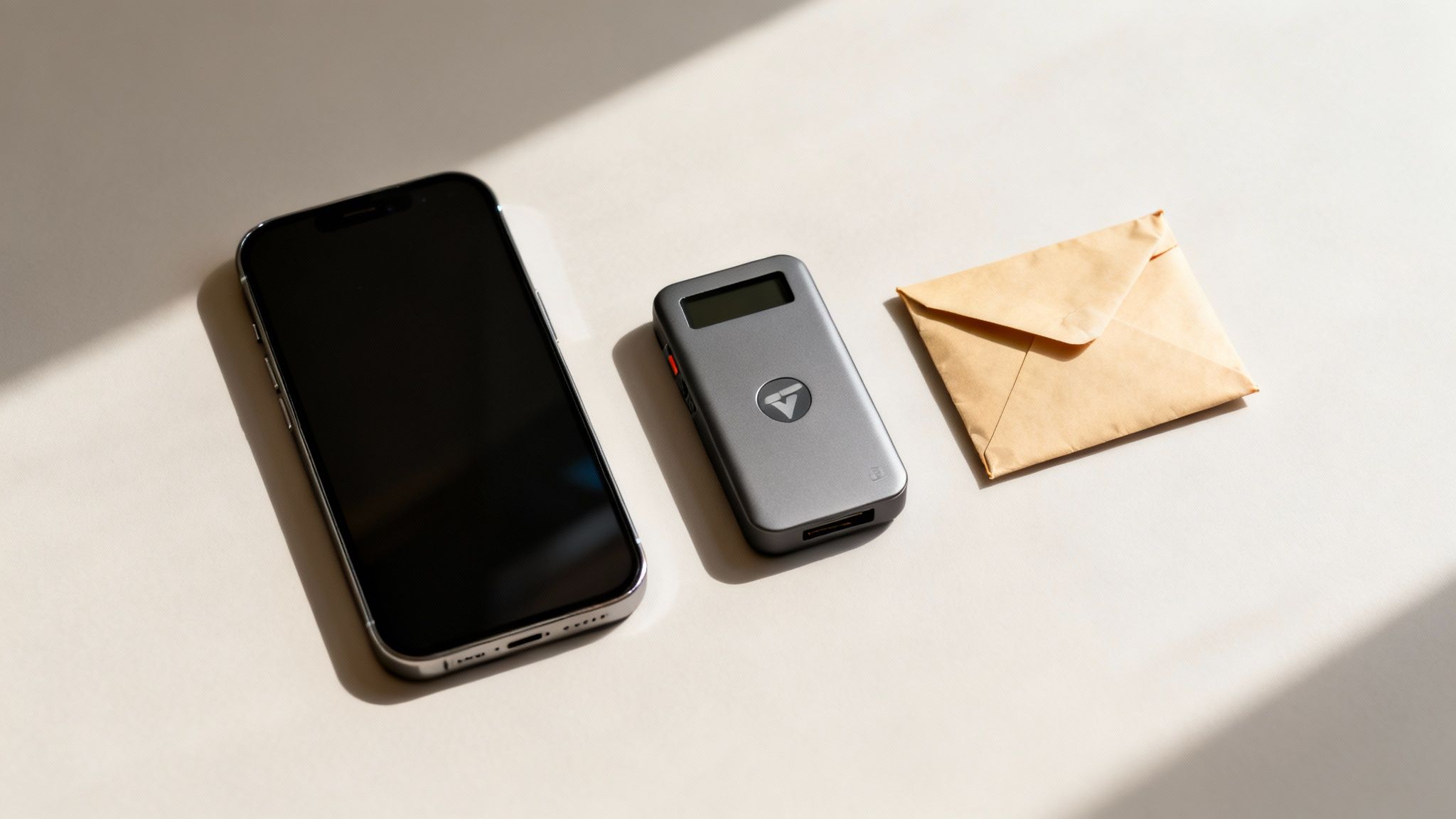 Ein Smartphone, ein graues Hardware-Wallet und ein brauner Umschlag auf hellem Hintergrund mit Schatten.