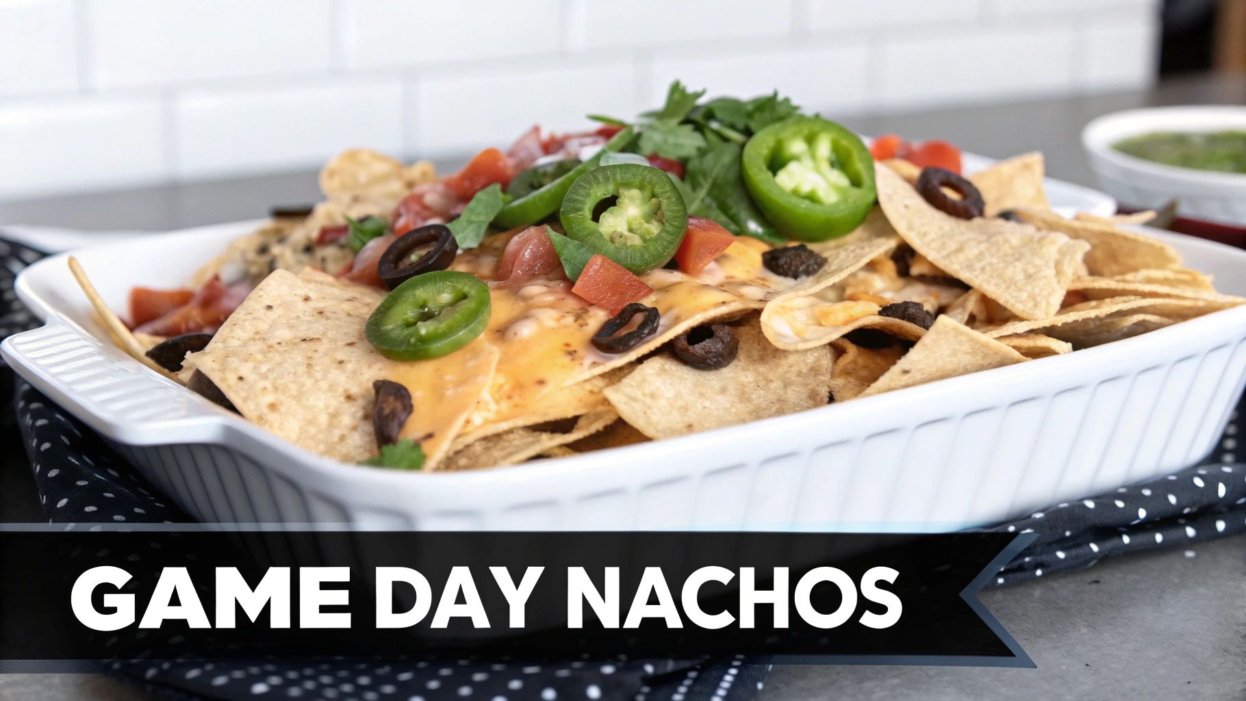 A white rectangular dish of game day nachos piled high with melted cheese, black olives, diced tomatoes, cilantro, and sliced jalapeños.