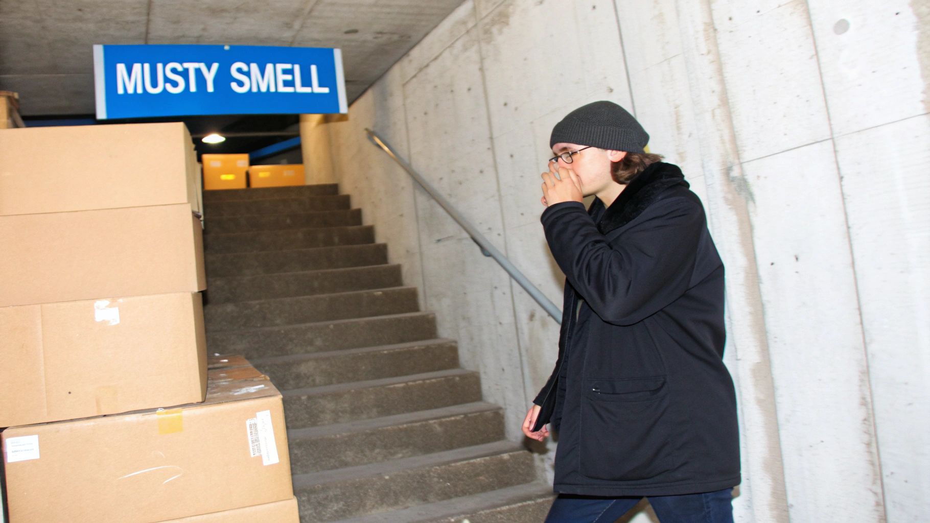 A person covers their nose near a "MUSTY SMELL" sign and stacked cardboard boxes on a concrete staircase.