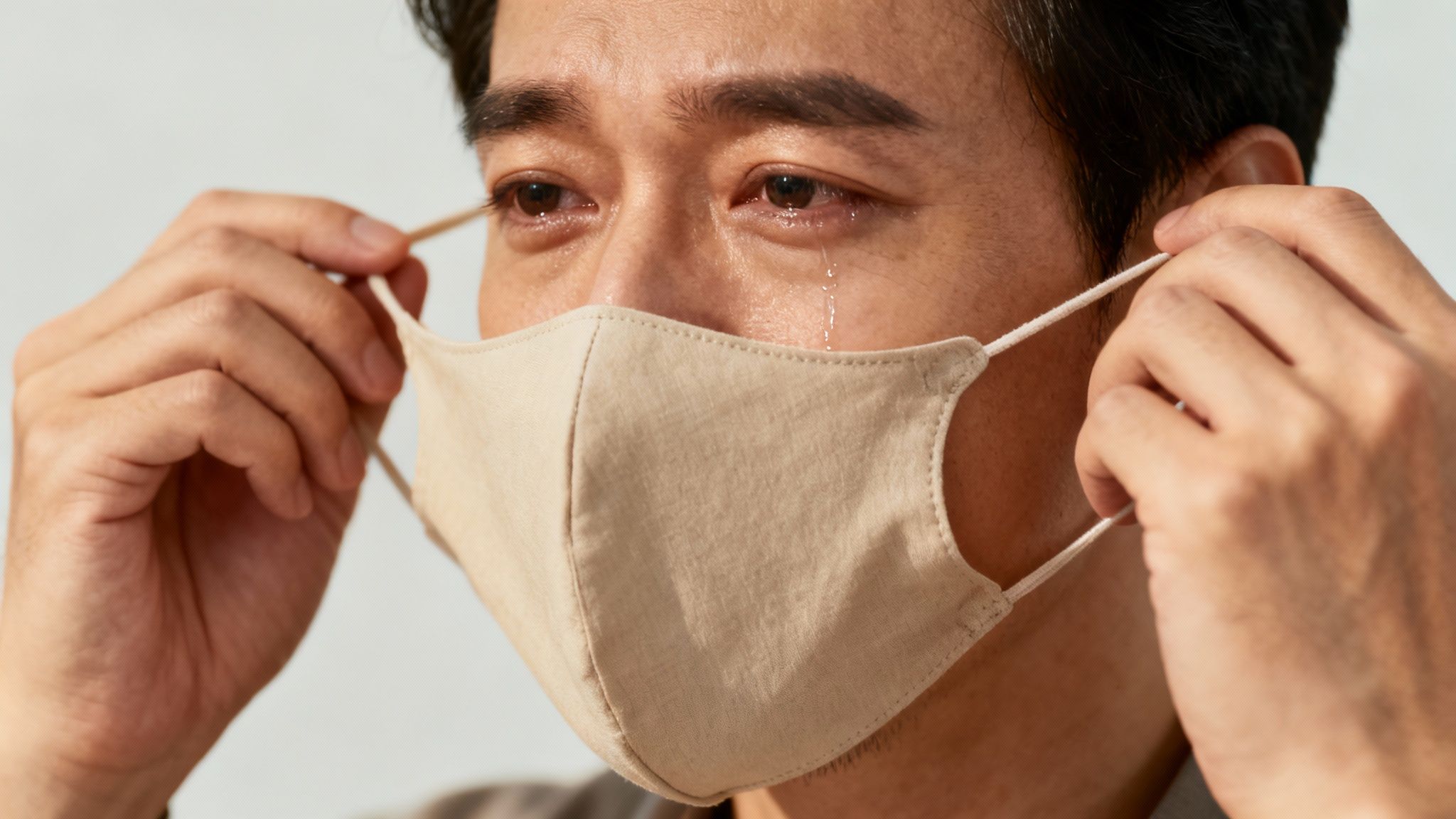 Close-up of an emotional man crying, holding a beige face mask near his face.
