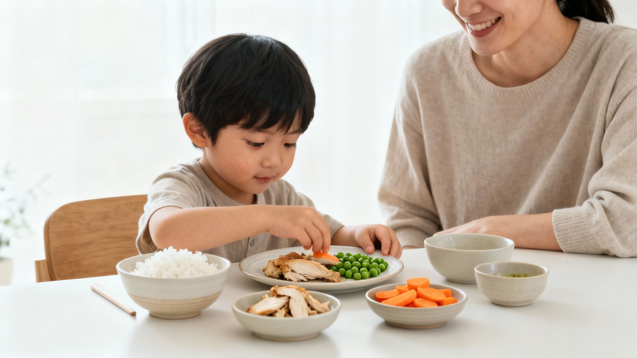A happy child picks up a carrot from a plate of food, with an adult smiling nearby during mealtime.
