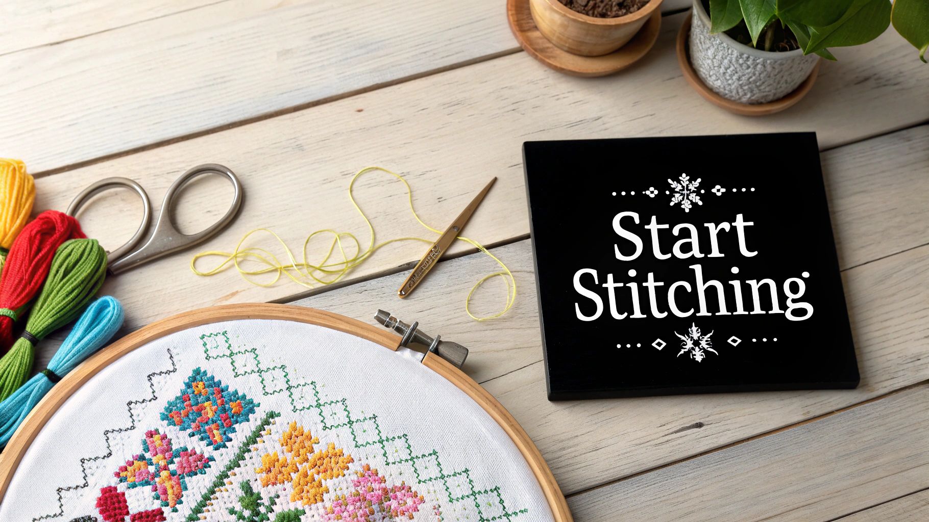 A flat lay of cross-stitching supplies including a hoop with a partially stitched pattern, colorful floss, and a 'Start Stitching' sign on a wooden table.