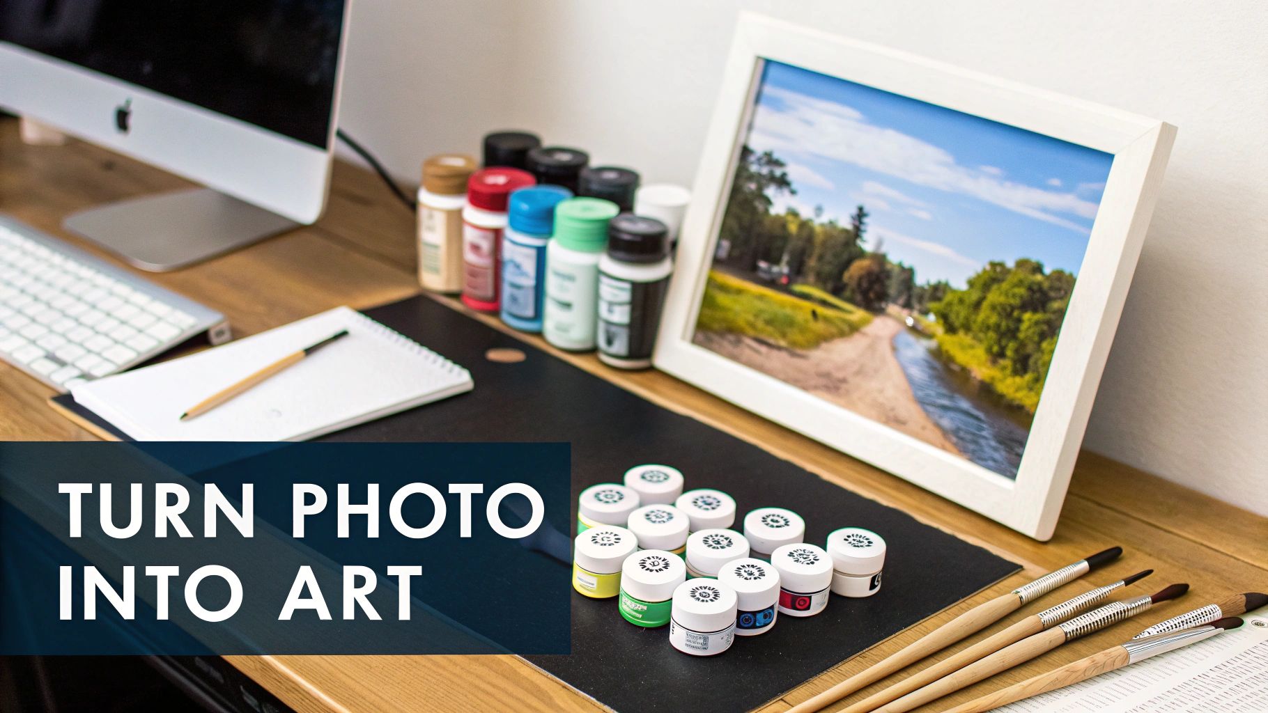 A creative workspace with a computer, a framed landscape photo, paint-by-numbers kit, and art brushes.