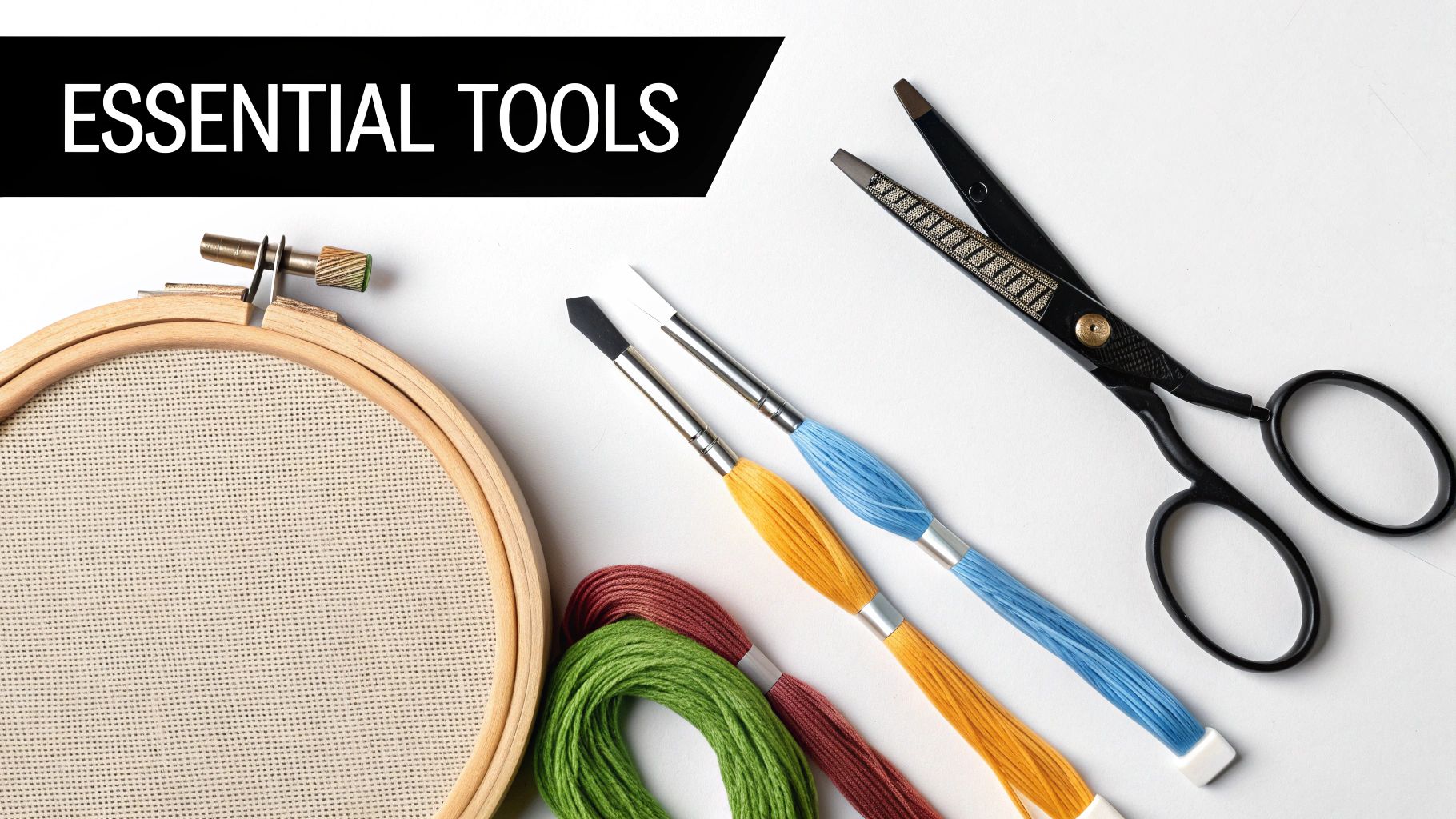 A flat lay of essential embroidery tools: an embroidery hoop with Aida fabric, colorful floss, brushes, and scissors.
