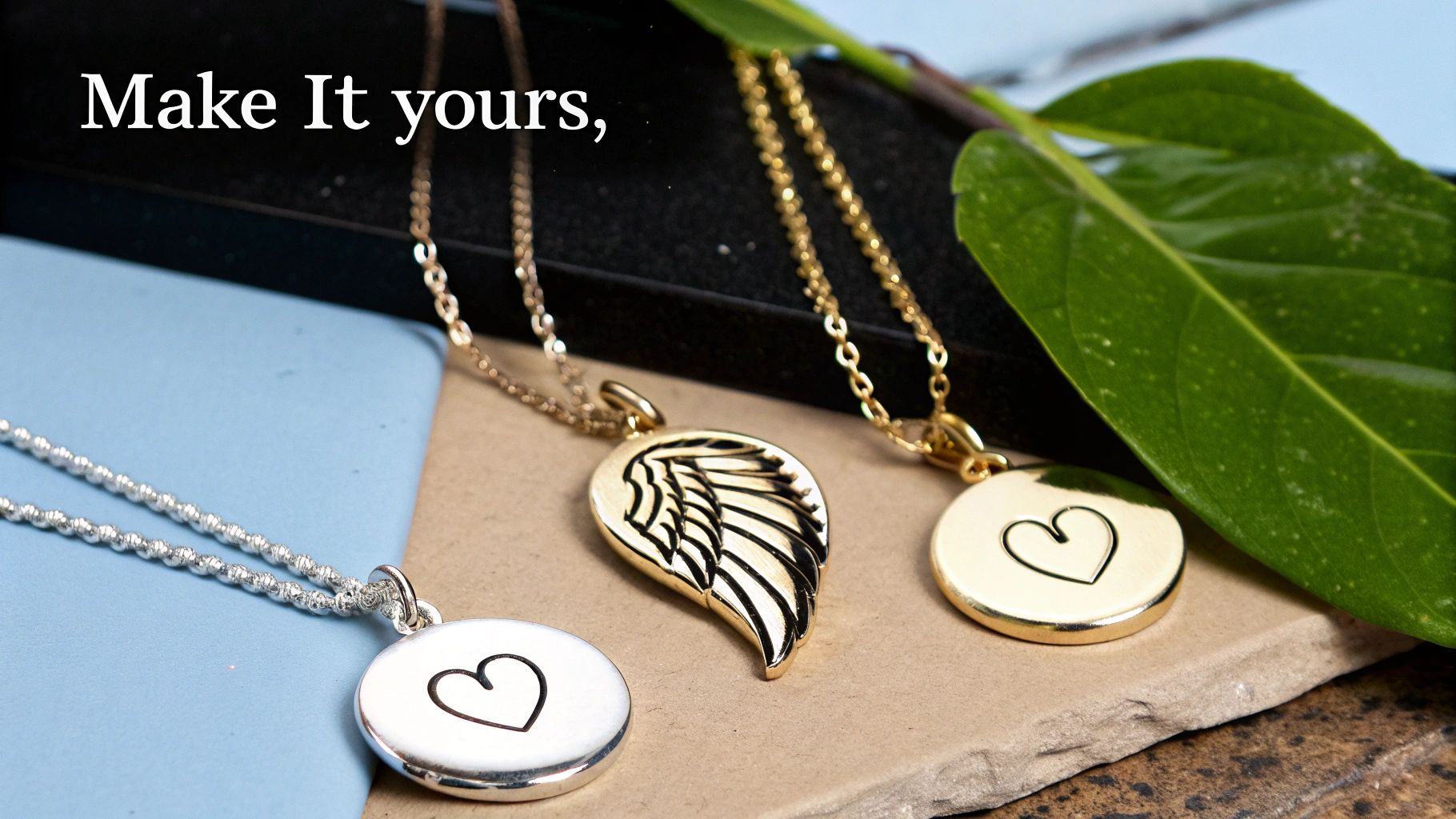 Three personalized necklaces, one silver heart, one gold wing, and one gold heart, with a green leaf.