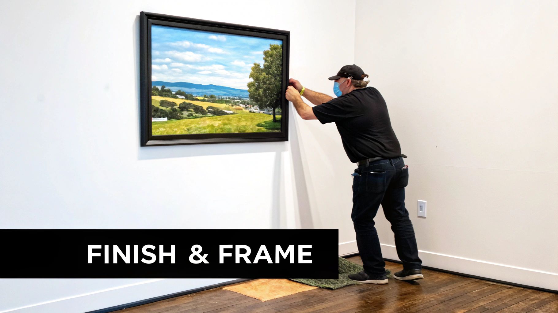 A person wearing a mask and cap hangs a framed landscape painting on a white wall.