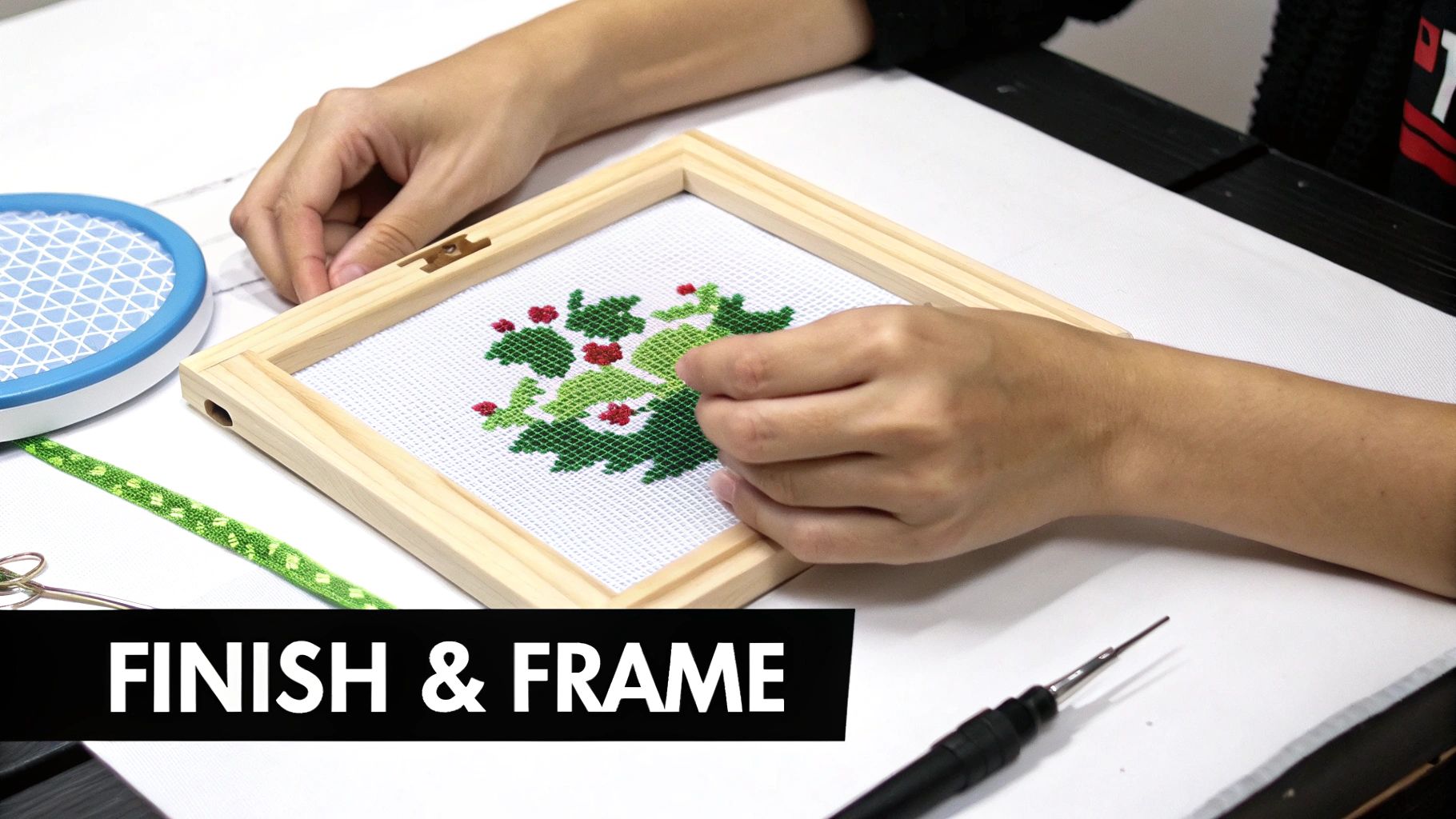 Close-up of hands cross-stitching a festive design with green leaves and red berries in a wooden frame, surrounded by crafting tools.