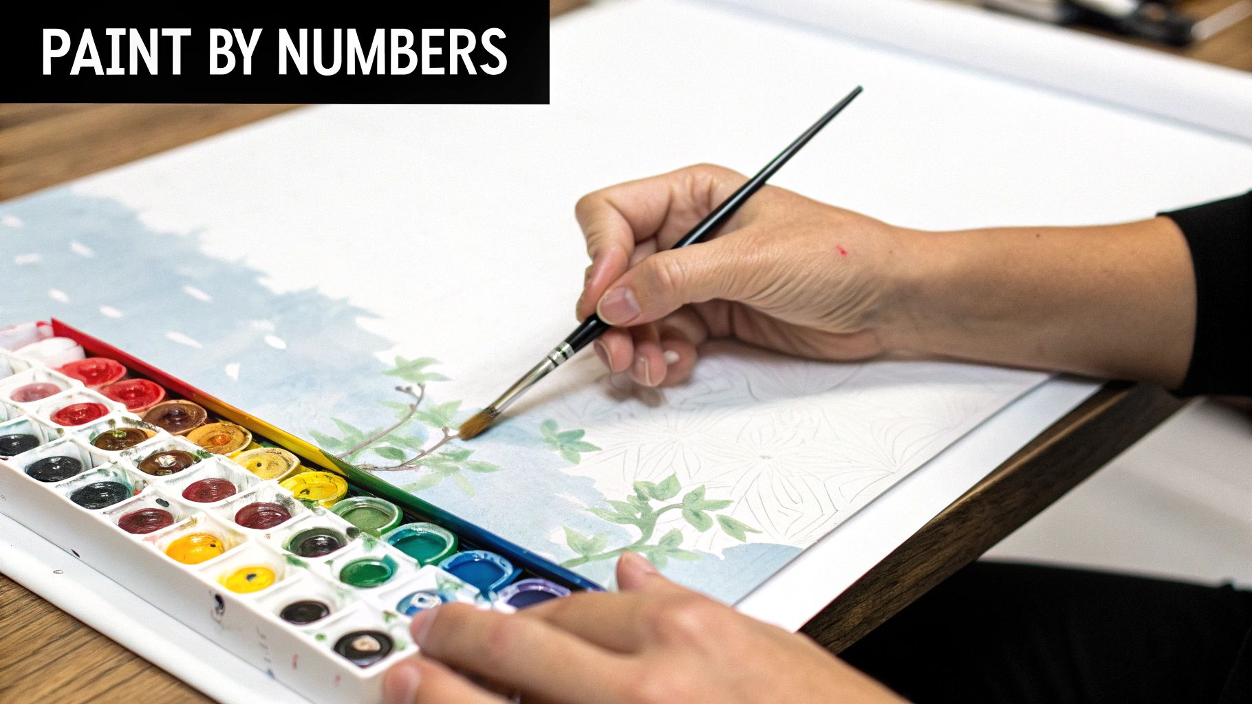 A person's hands paint a nature scene with watercolors from a palette on a paint by numbers guide.