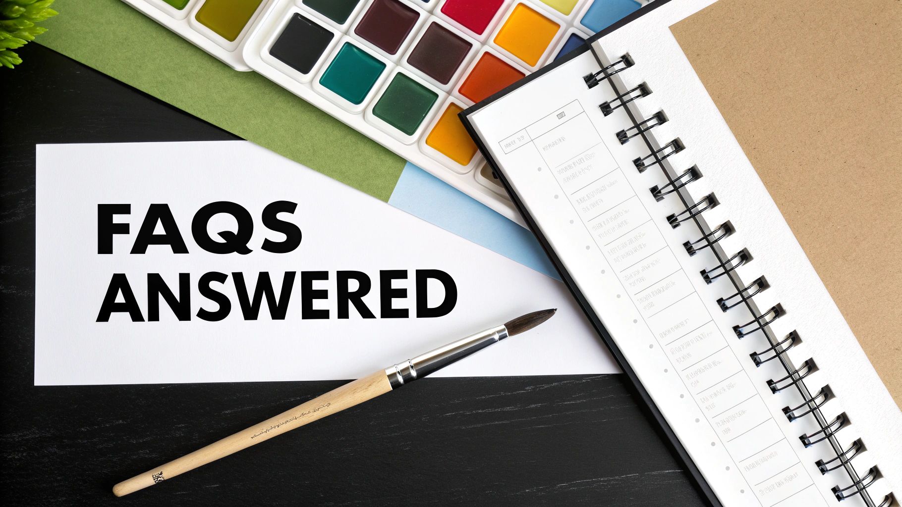 White paper with 'FAQS ANSWERED' text, a paintbrush, watercolor palette, and a spiral notebook on a desk.