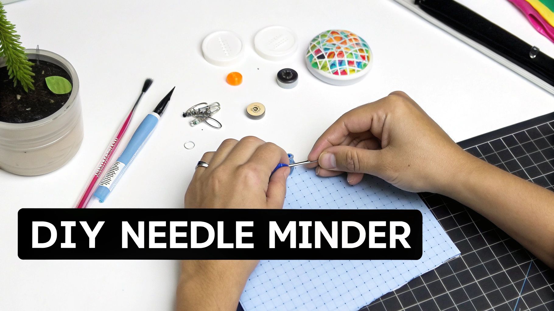 Hands crafting a colorful DIY needle minder from supplies on a white table with a plant.