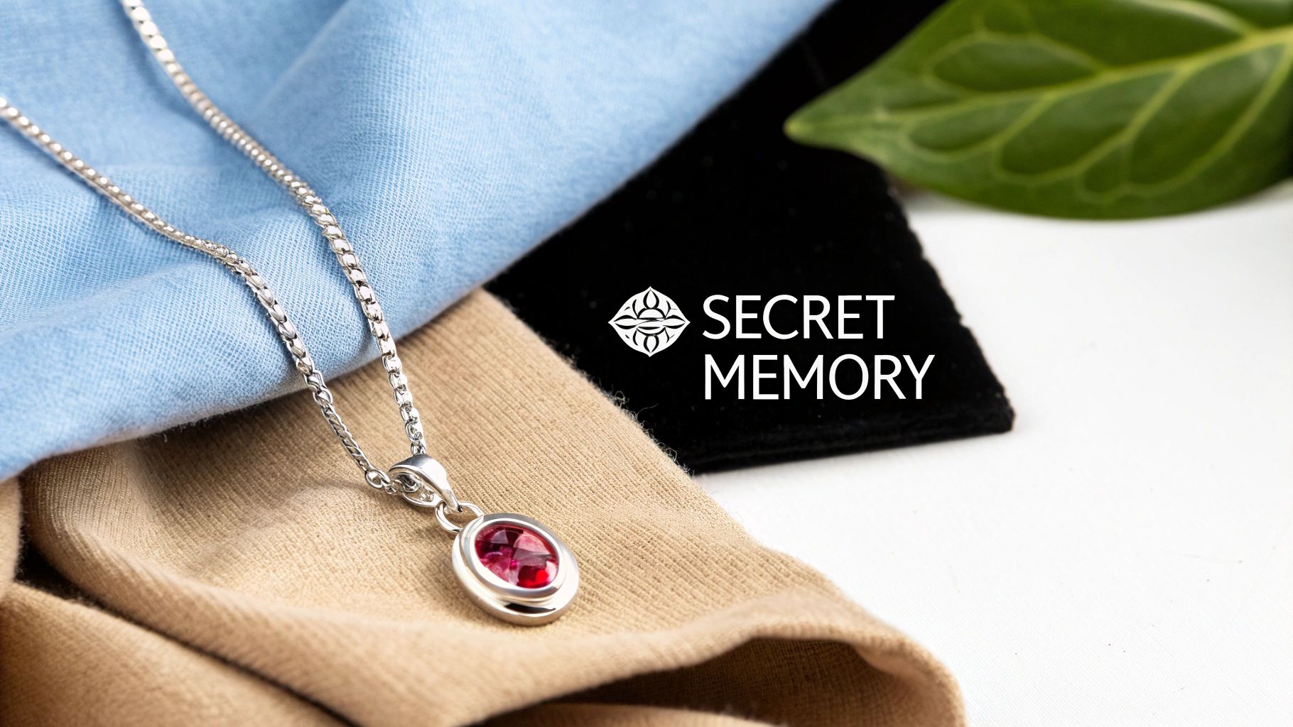 A silver necklace with a red oval gem pendant rests on light blue and beige fabrics, with a 'SECRET MEMORY' logo.