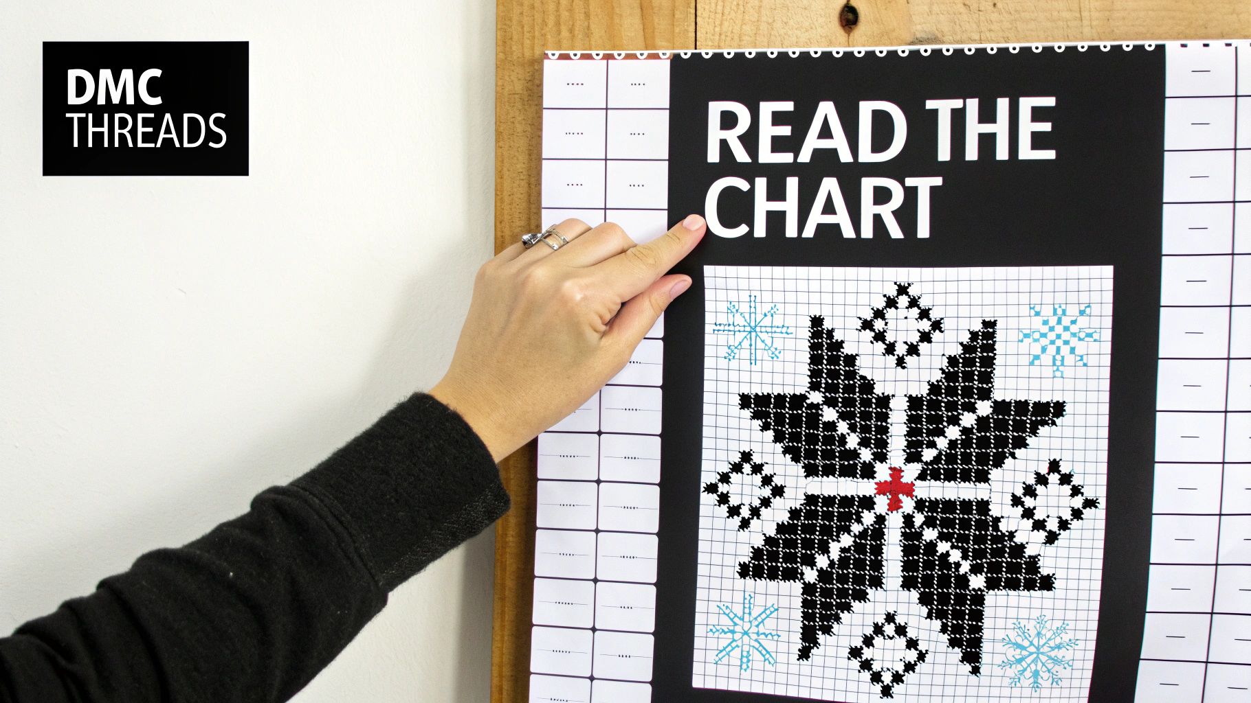 A hand points to a DMC Threads cross-stitch chart displaying a black, red, and blue snowflake design.