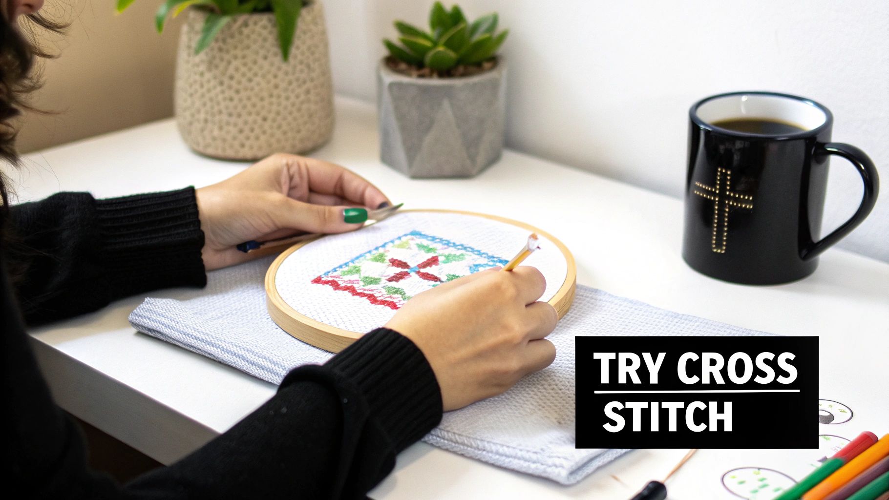 Close-up of hands working on a colorful cross-stitch project in a hoop on a white desk, with plants and a coffee mug.